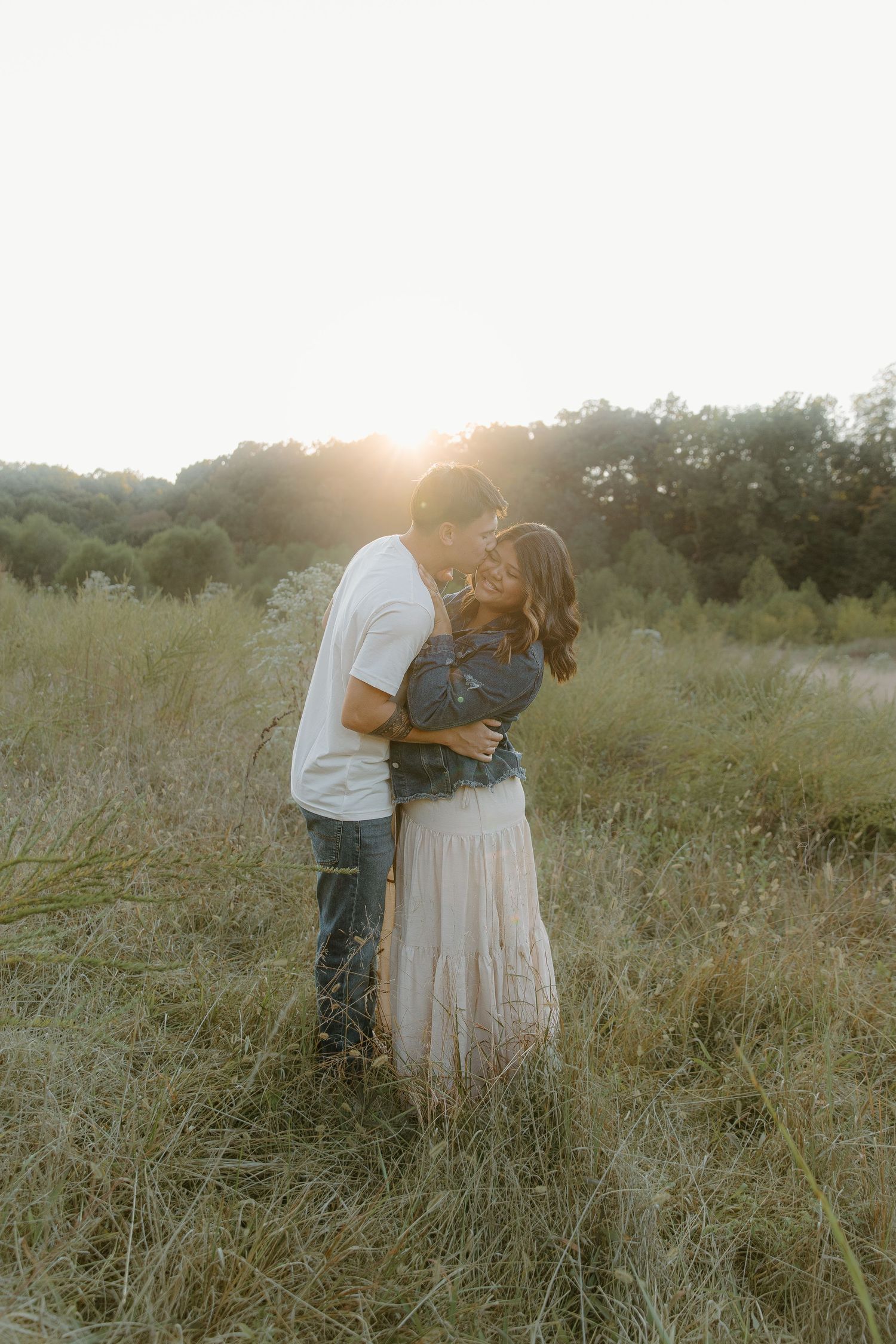 Couple embraces and kisses in a golden sunlit meadow field during a romantic outdoor engagement session.