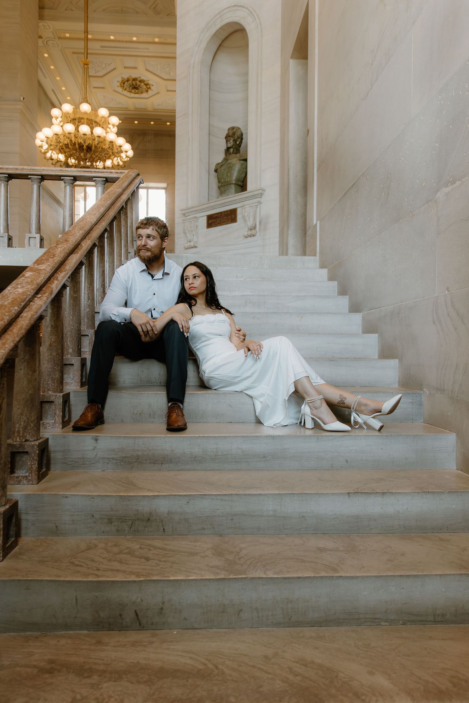 Bride in white dress and groom sit relaxed on marble stairs in elegant historic building.
