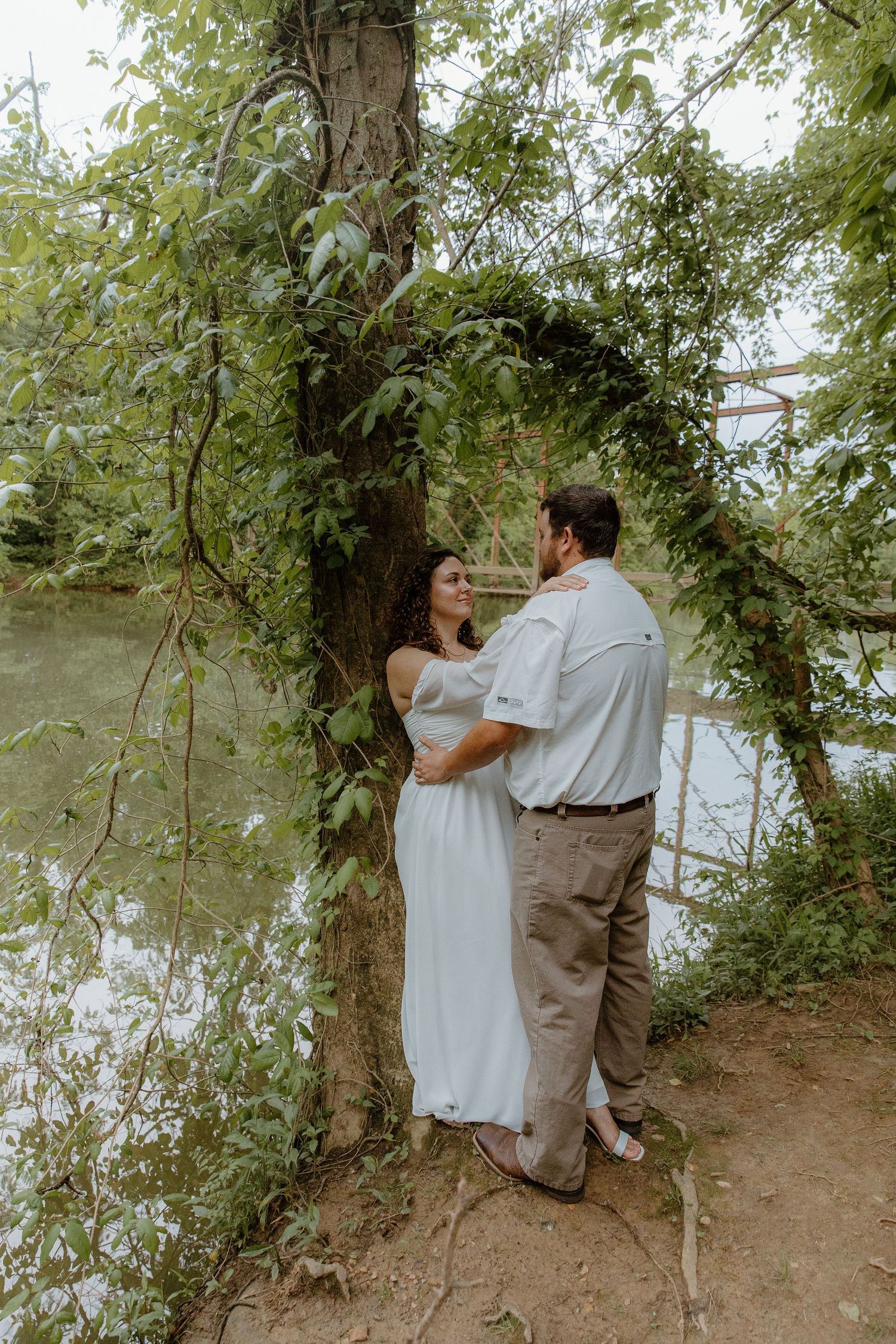 Couple embracing under a lush green tree arch near a serene river, woman in white dress, man in casual attire.