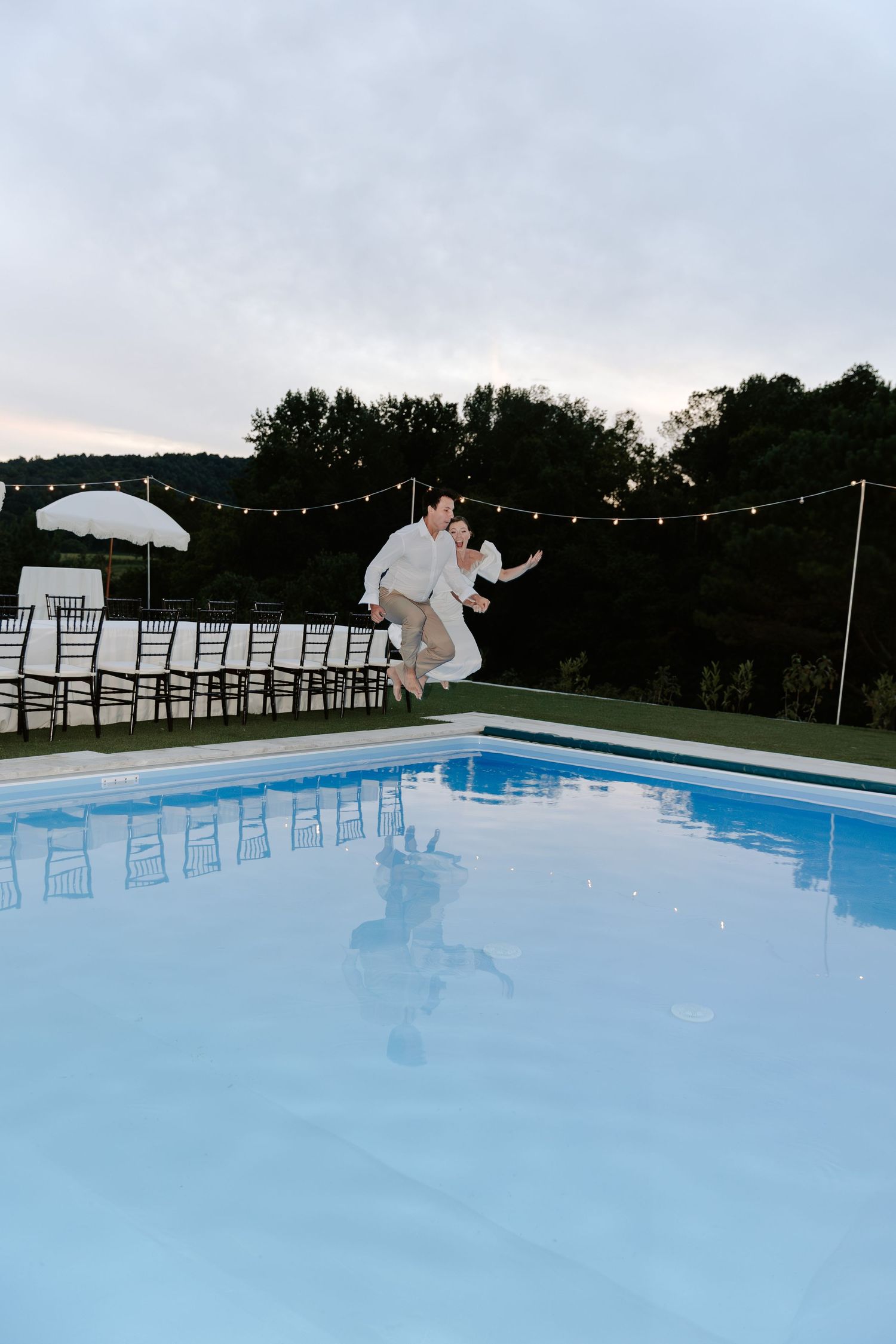 Bride and groom jumping into a pool at dusk with string lights in background.