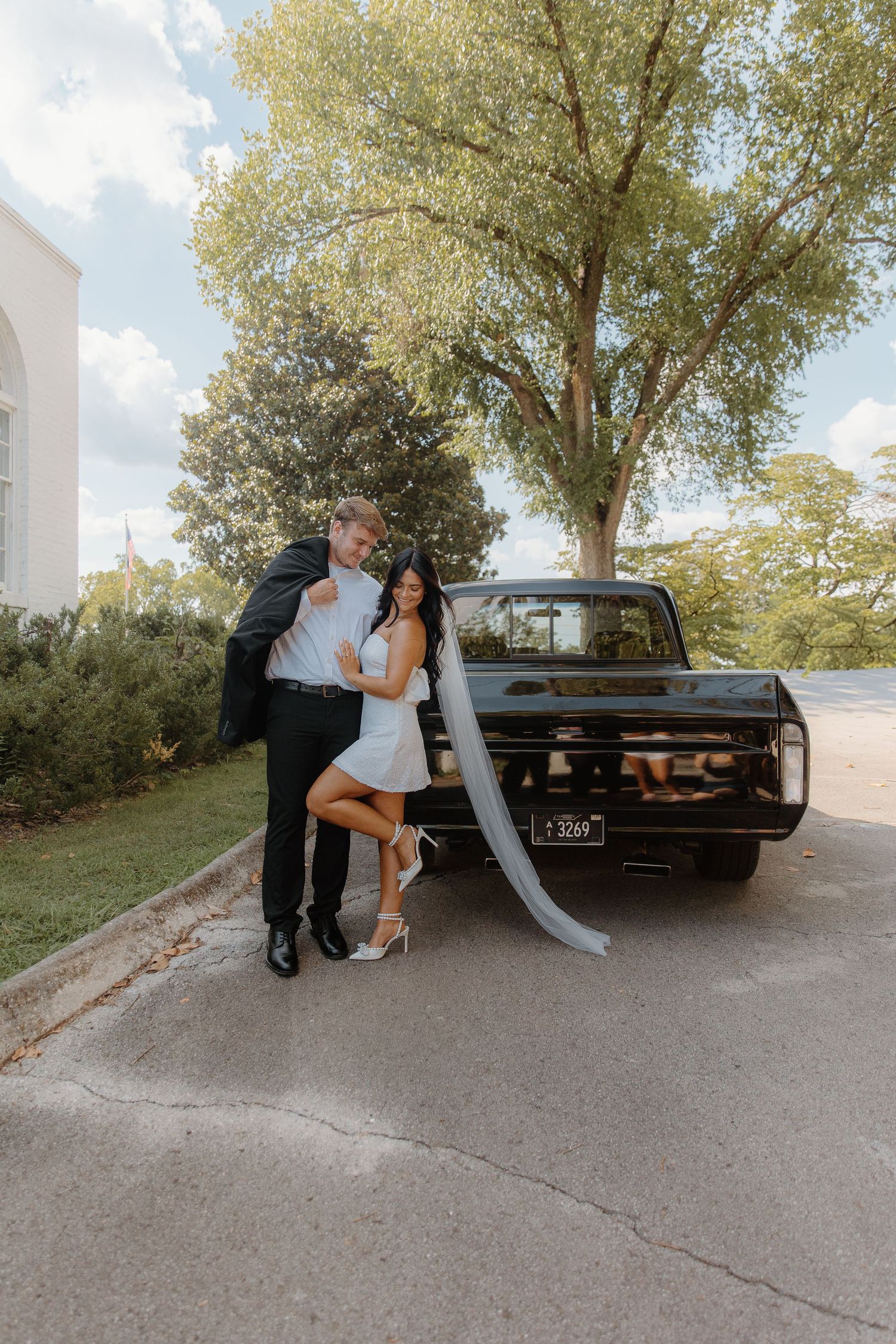 Bride in short white dress and groom pose beside a vintage black truck under large trees.