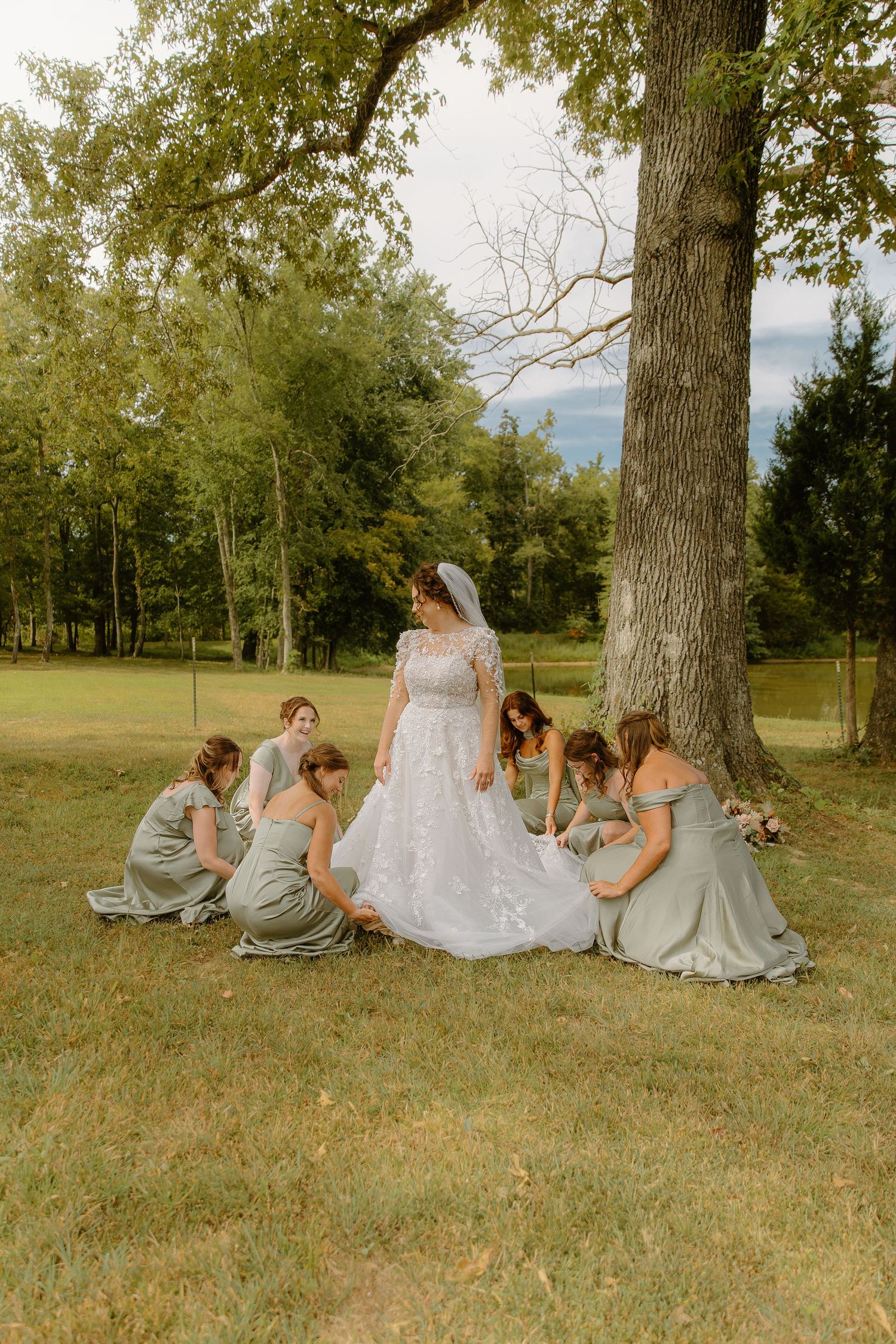 Bride in white lace gown with bridesmaids in sage green dresses arranging her train under a large tree outdoors.