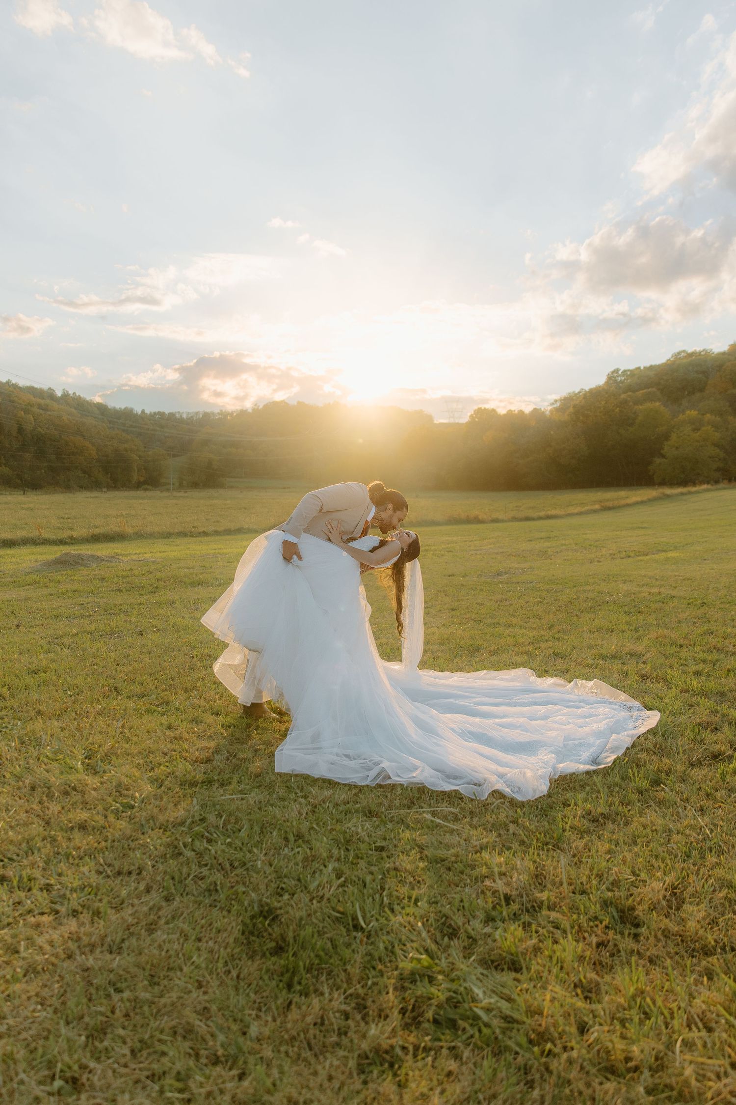 Bride and groom share a romantic dip kiss at golden sunset in a lush green meadow with rolling hills.
