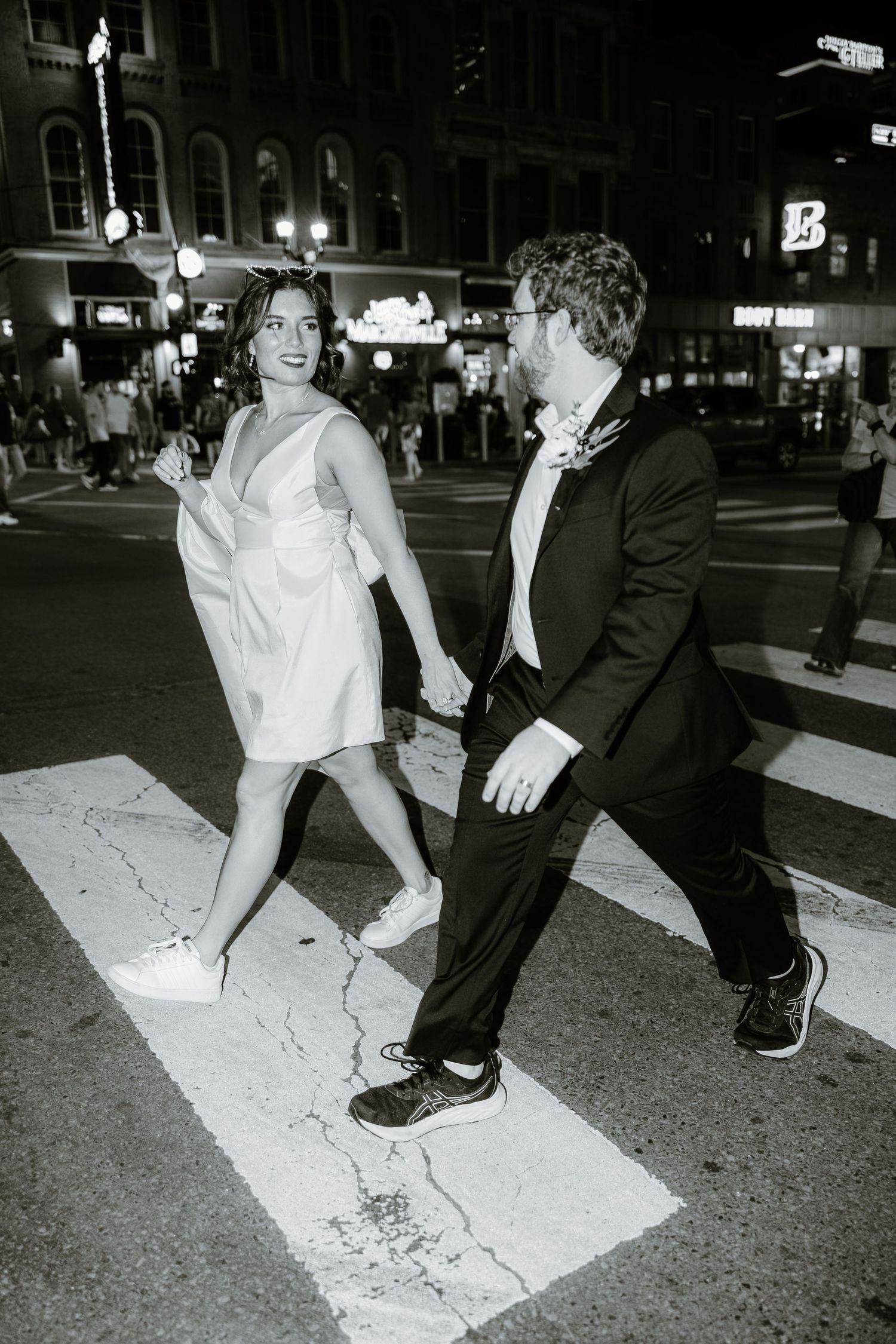 Bride and groom crossing a city street at night, she in white dress and sneakers, he in black suit.