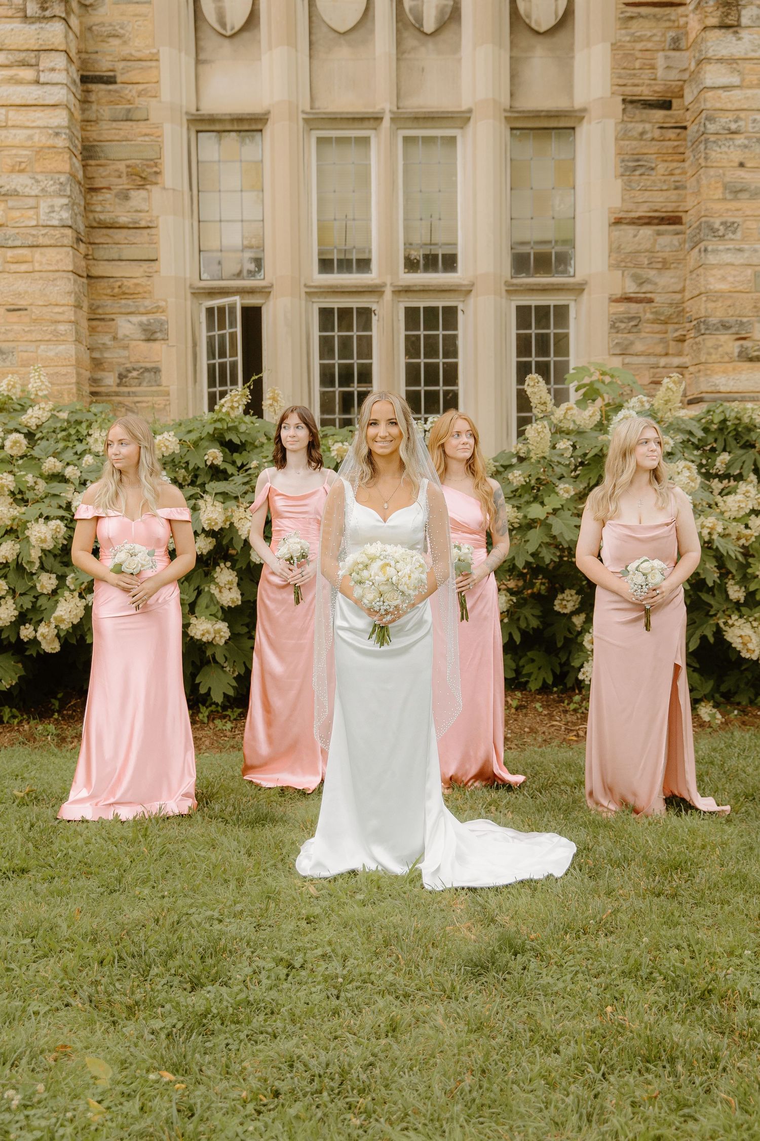 Bride in white gown stands with four bridesmaids in pink satin dresses in front of a stone building.