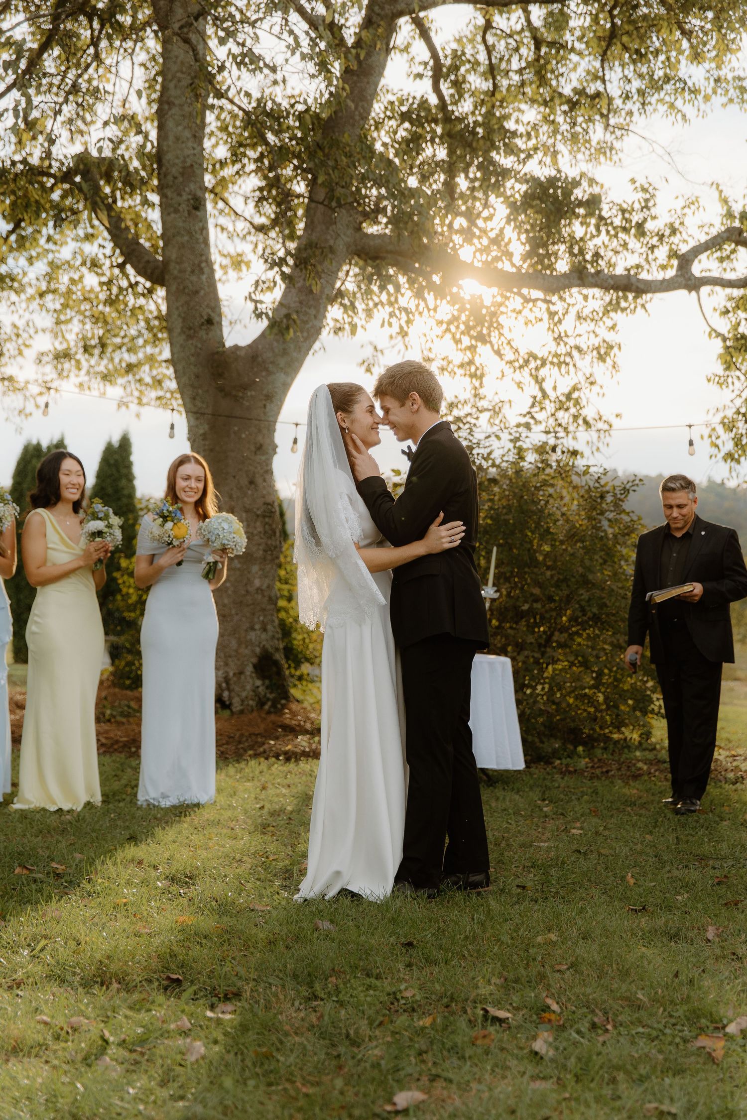 Bride and groom share first kiss under a sunlit tree during outdoor wedding ceremony with bridesmaids nearby.