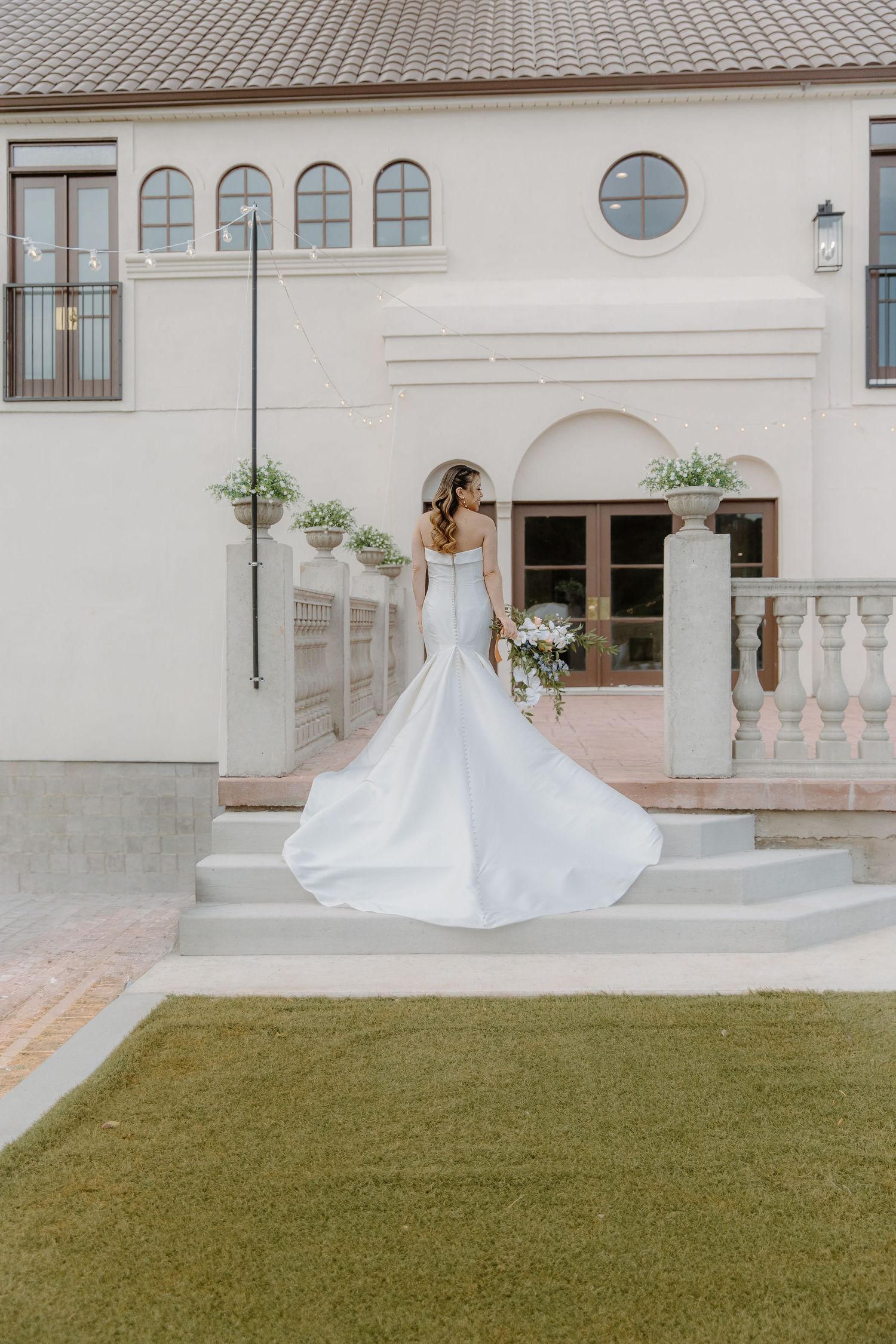 Bride in elegant white mermaid wedding gown with flowing train stands on steps outside a beautiful venue.