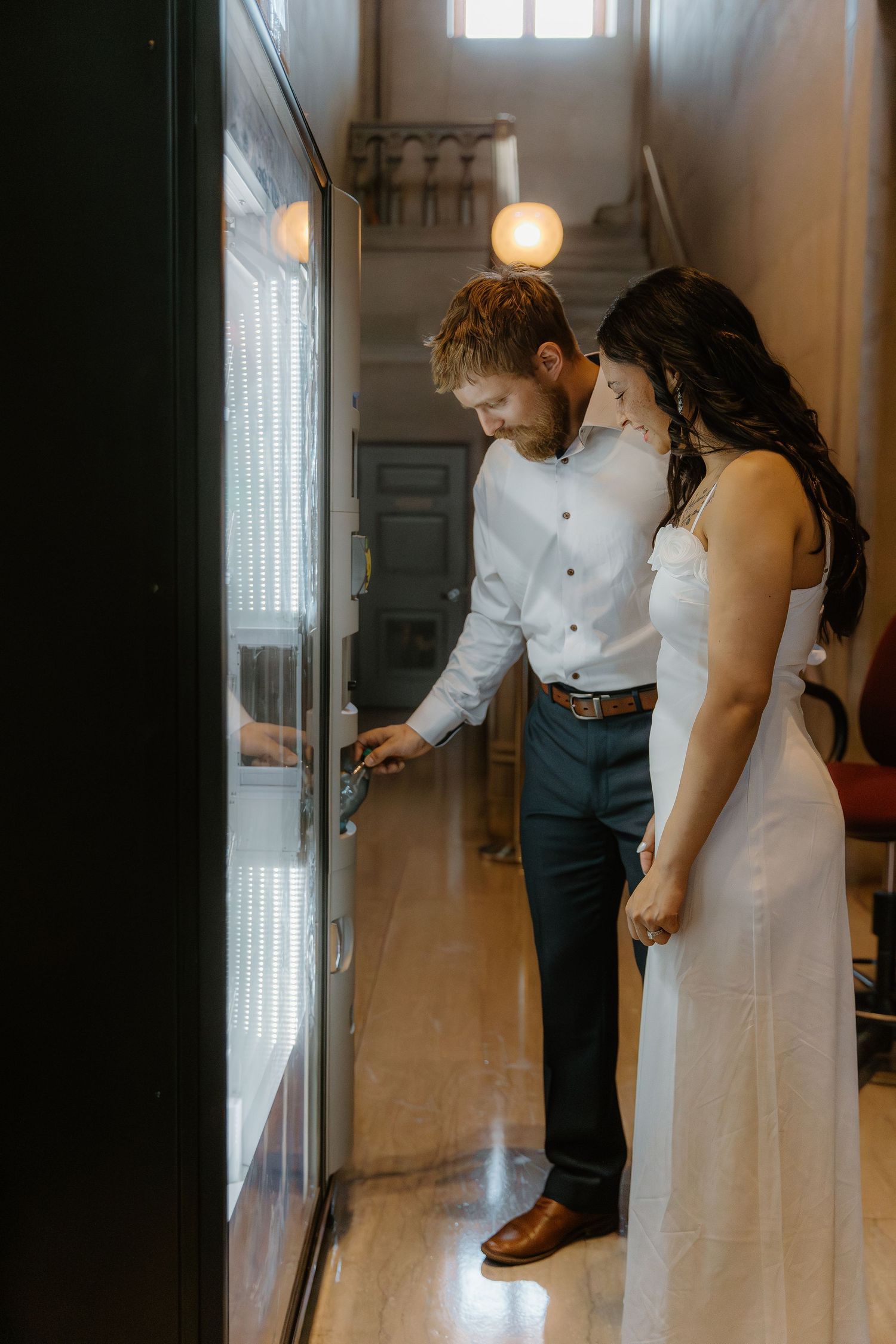 Bride and groom in casual wedding attire browse a vending machine together in a warmly lit hallway.