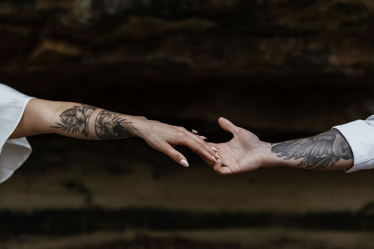 Two tattooed hands reaching toward each other with an engagement ring visible.