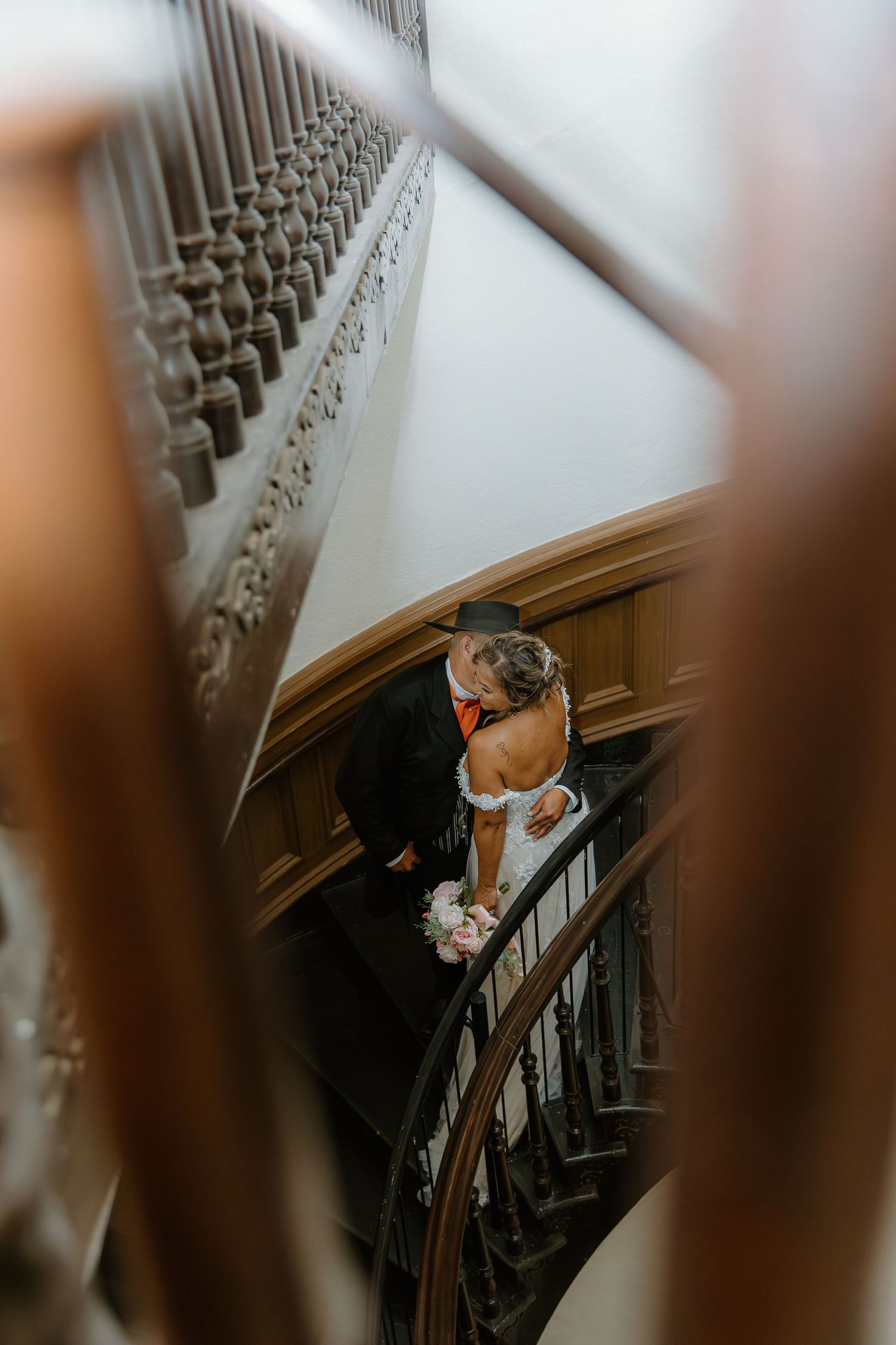 Bride and groom sharing a romantic kiss on an ornate wooden staircase, captured from above during their wedding day.