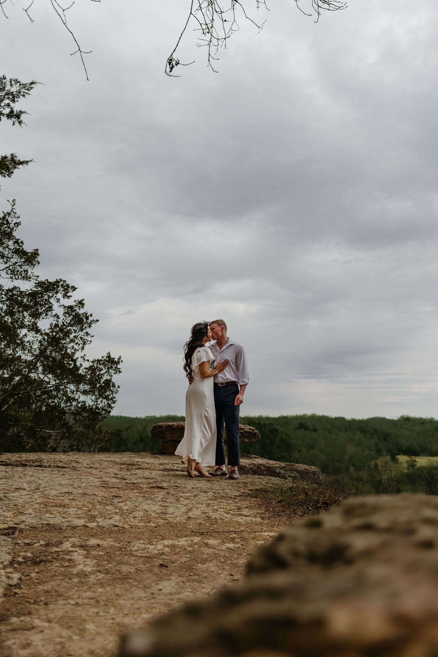 Couple sharing a romantic kiss on a rocky overlook surrounded by lush green trees under a cloudy sky.