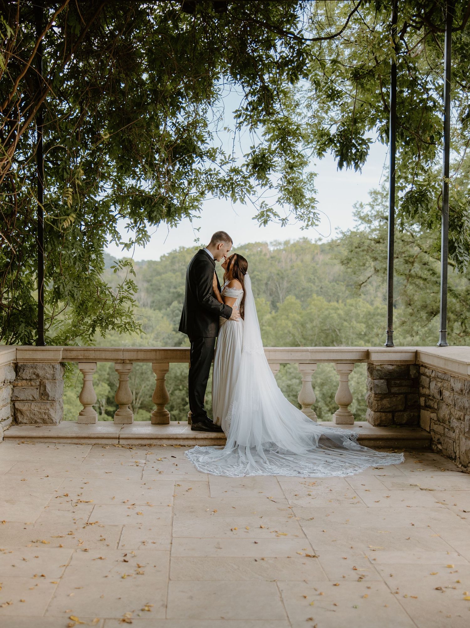 Bride and groom share a romantic moment on a stone terrace balcony surrounded by lush green trees.