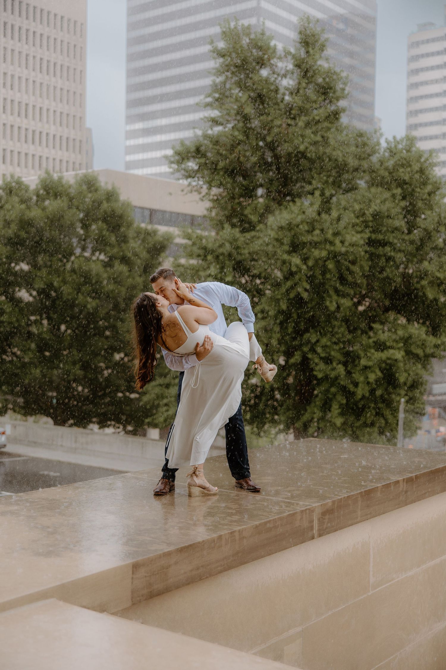 Couple sharing a romantic kiss in the rain, with lush green trees and city skyscrapers in the background.