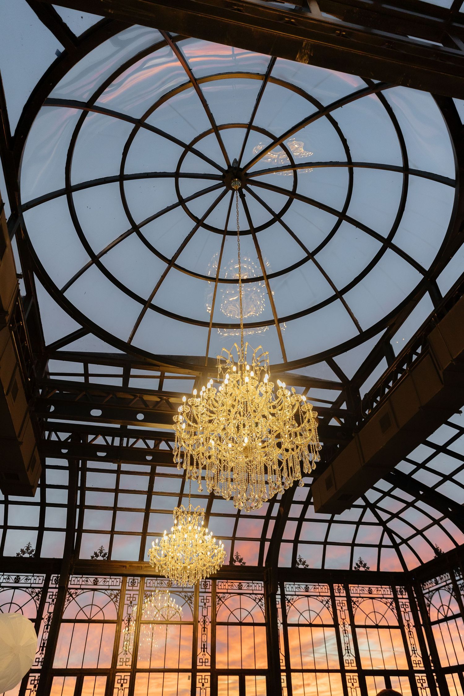 Elegant crystal chandeliers hanging beneath a circular glass dome ceiling at sunset.