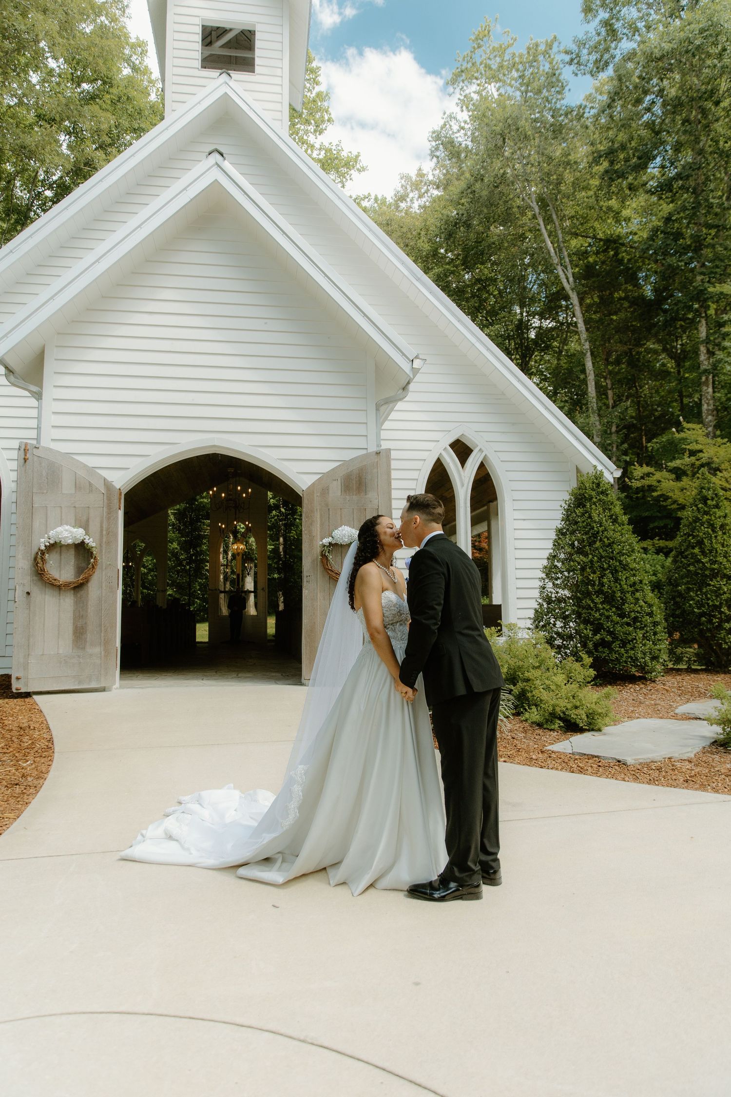 Bride and groom share a kiss outside a white chapel surrounded by lush trees on their wedding day.