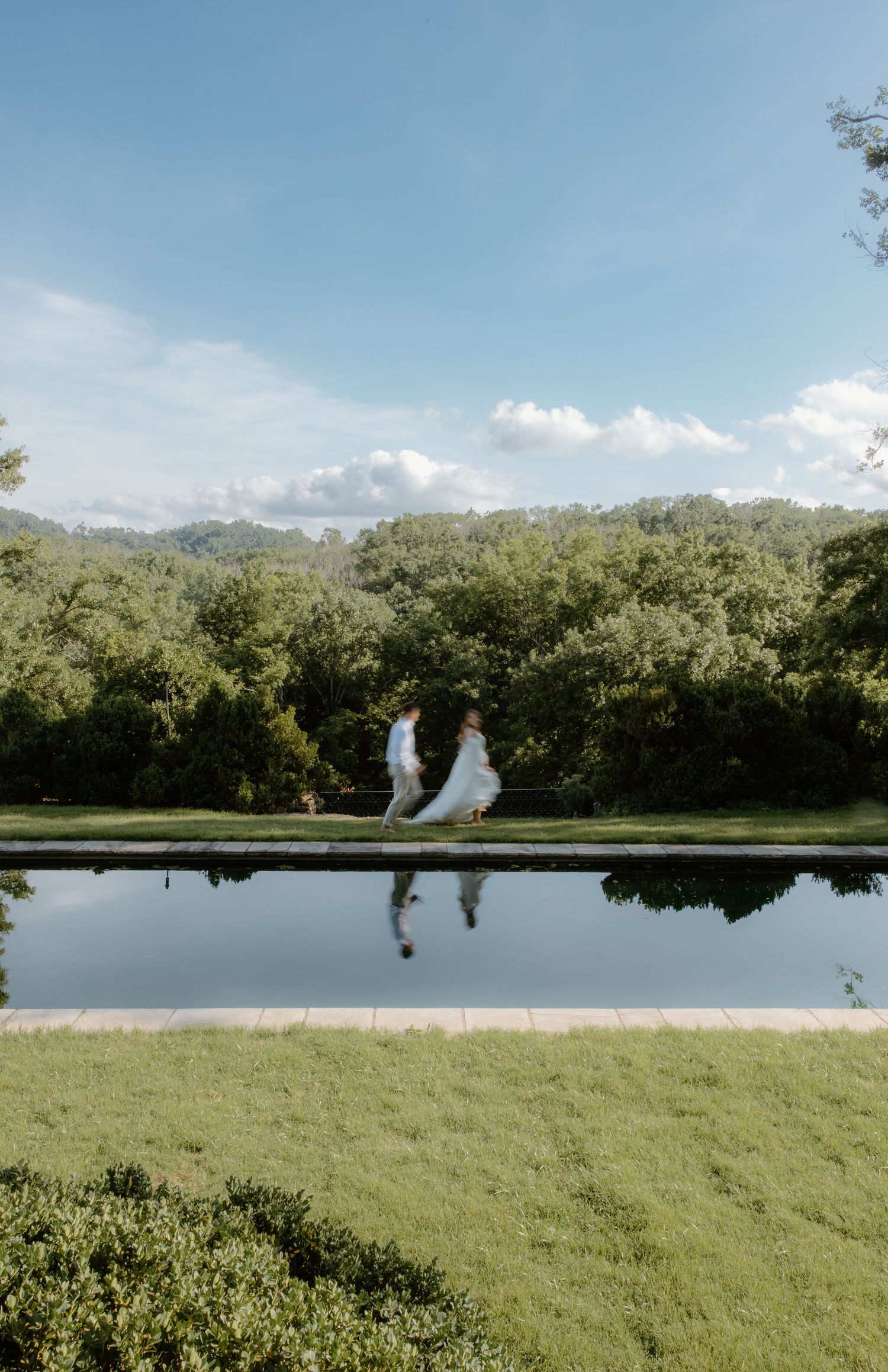 A bride and groom walk hand in hand beside a reflective pool surrounded by lush green trees on a sunny day.