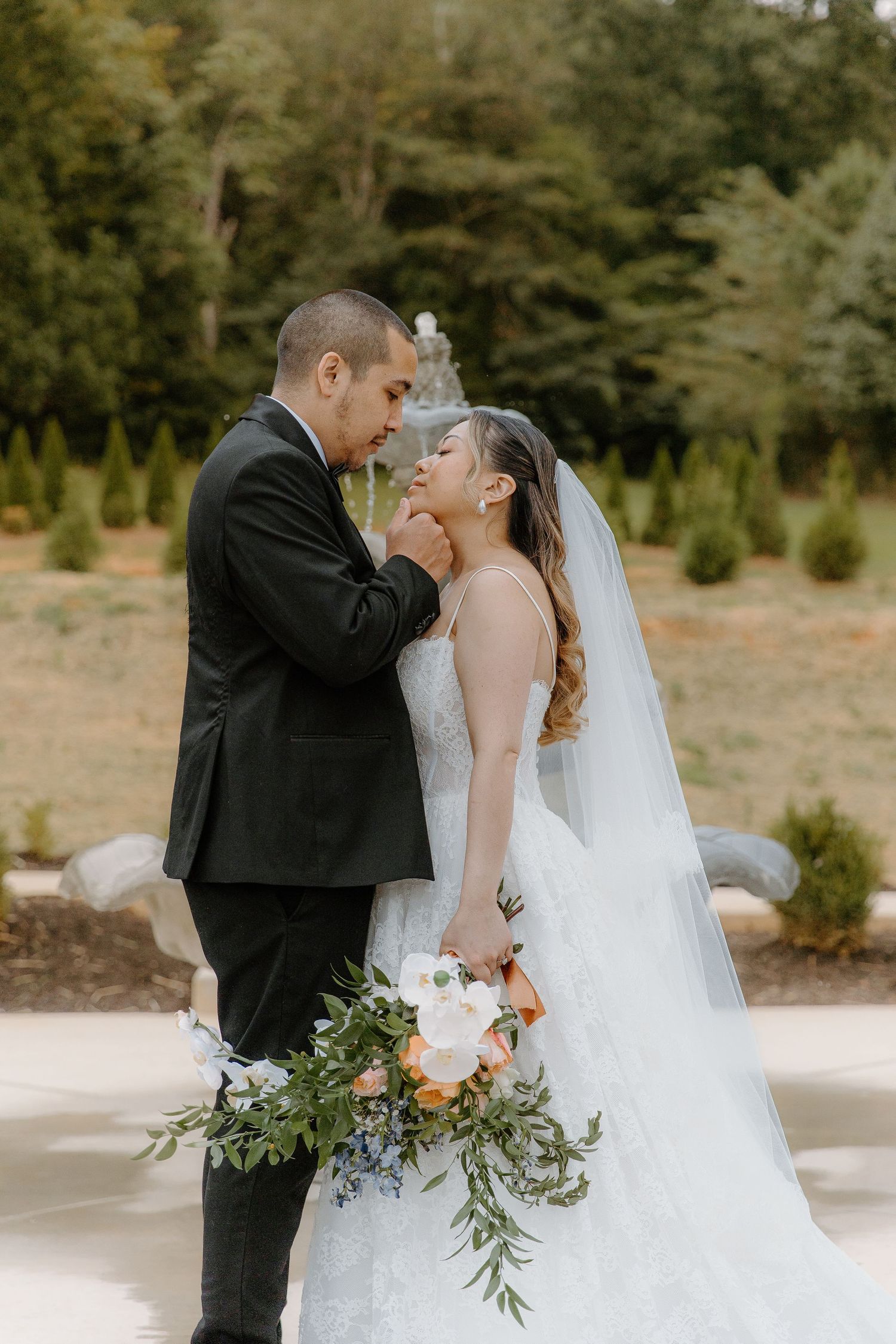 Bride and groom share an intimate moment outdoors, with the bride holding a lush bouquet of white and peach flowers.
