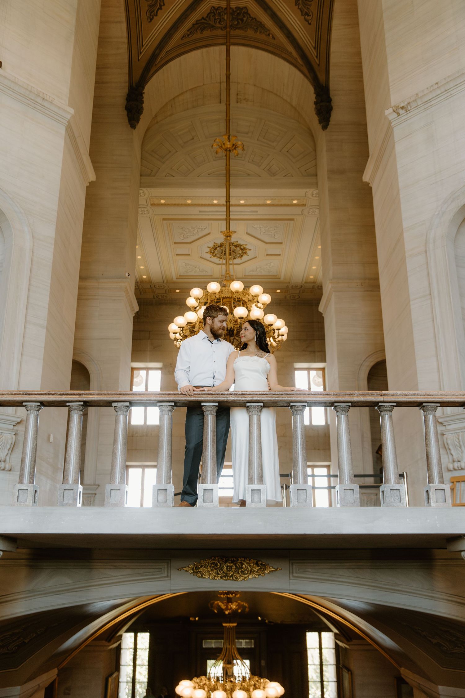 Bride and groom stand on a grand balcony inside an ornate historic building with gold chandelier.