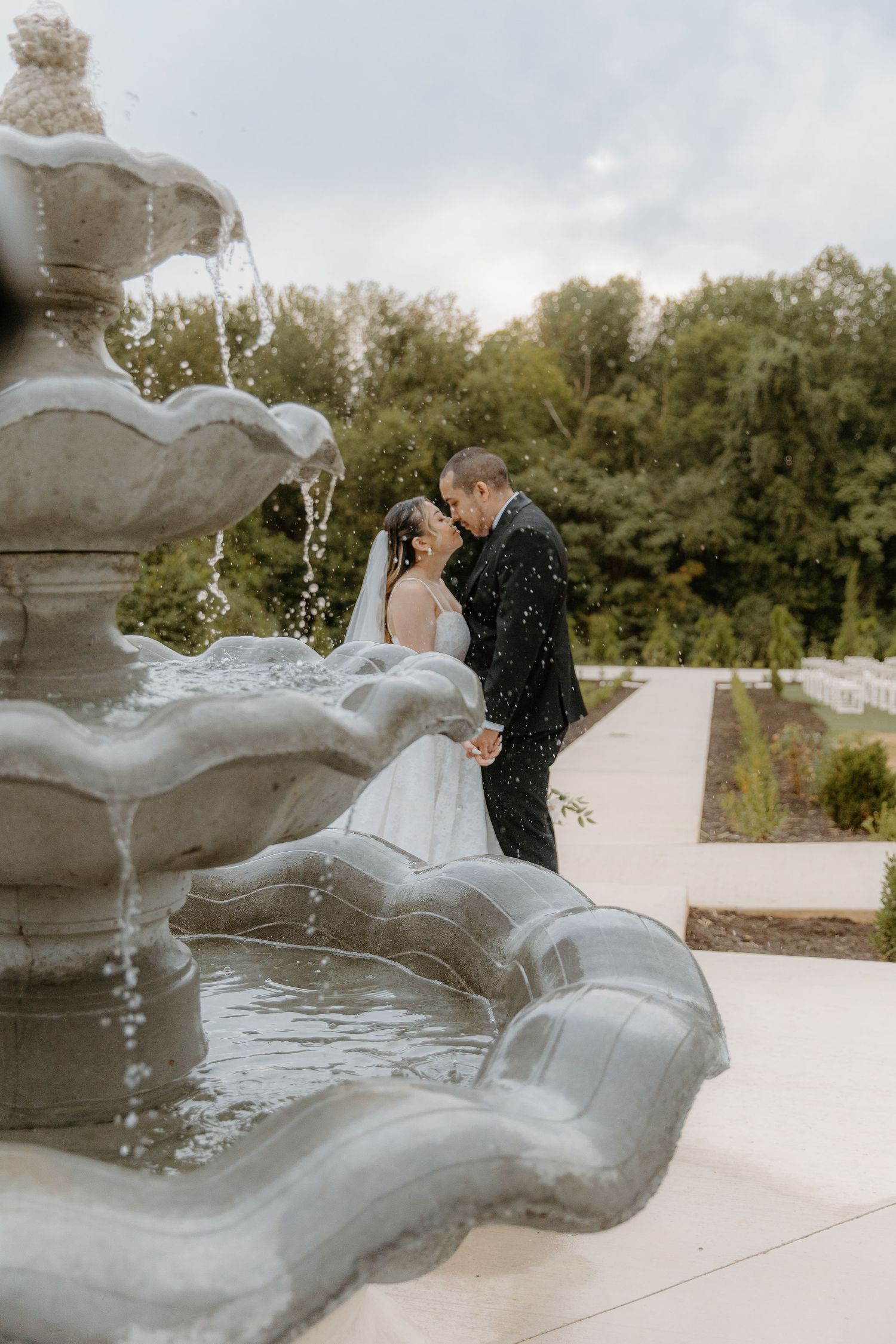 Bride and groom share a romantic kiss near an elegant stone fountain in a lush garden on their wedding day.