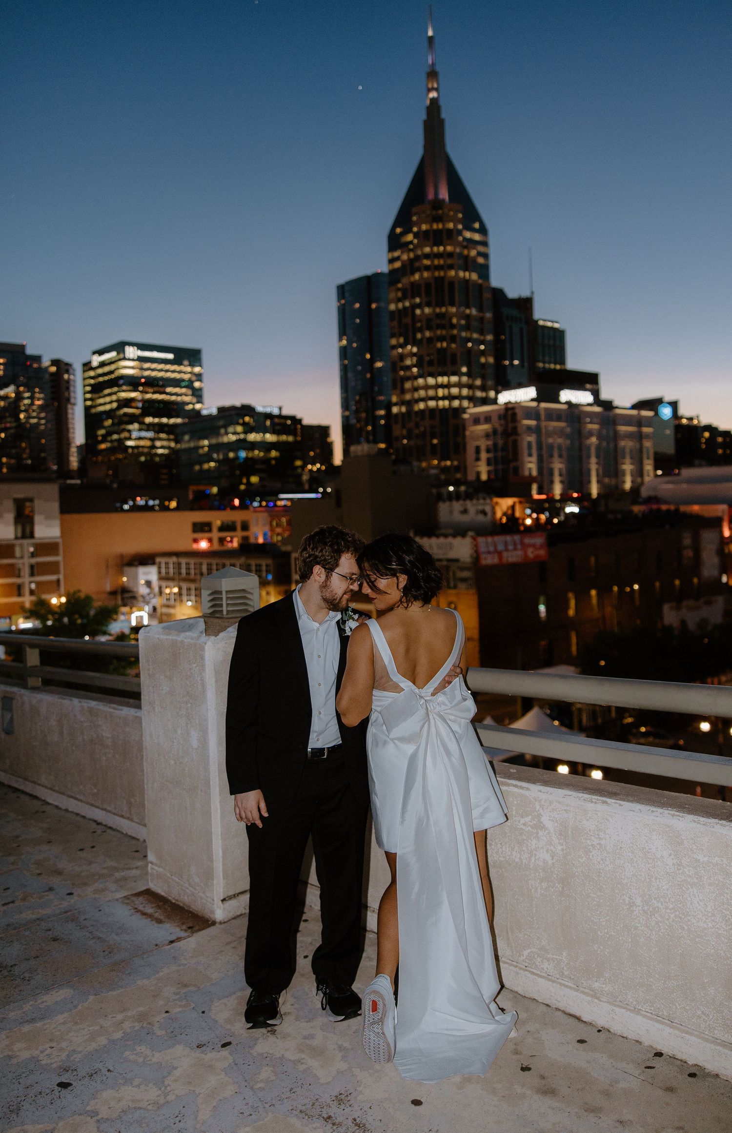 Couple embraces on rooftop at dusk with Nashville city skyline, bride in backless bow gown, groom in black suit.