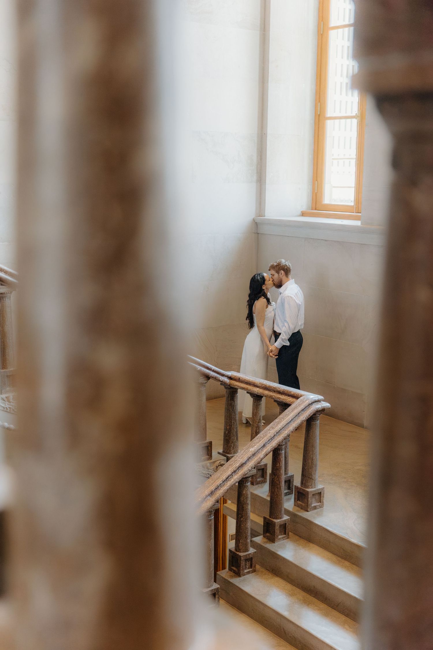 Couple sharing a romantic kiss on elegant wooden staircase in a sunlit historic building.