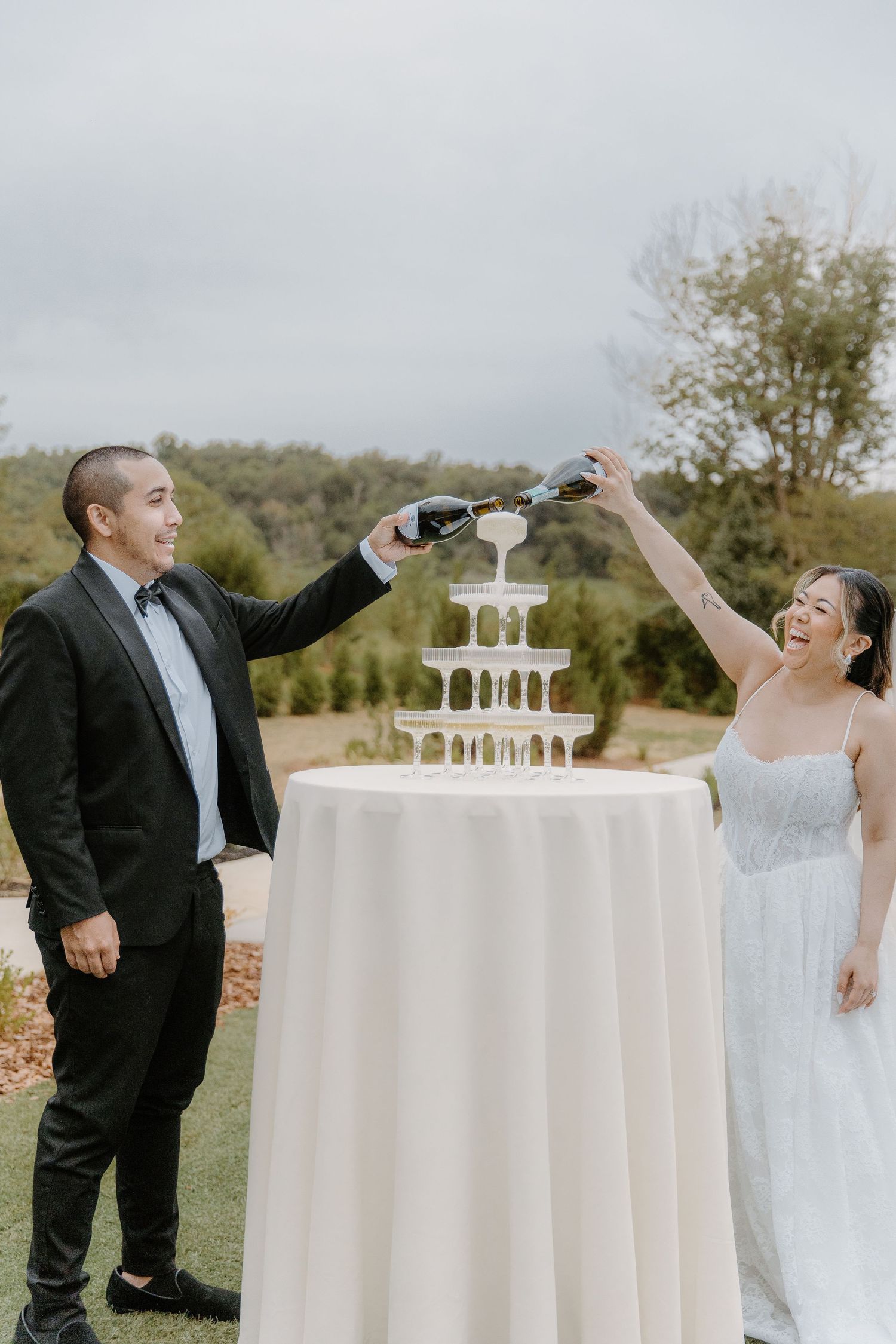 Bride and groom joyfully pouring champagne into a tower at an outdoor wedding celebration.