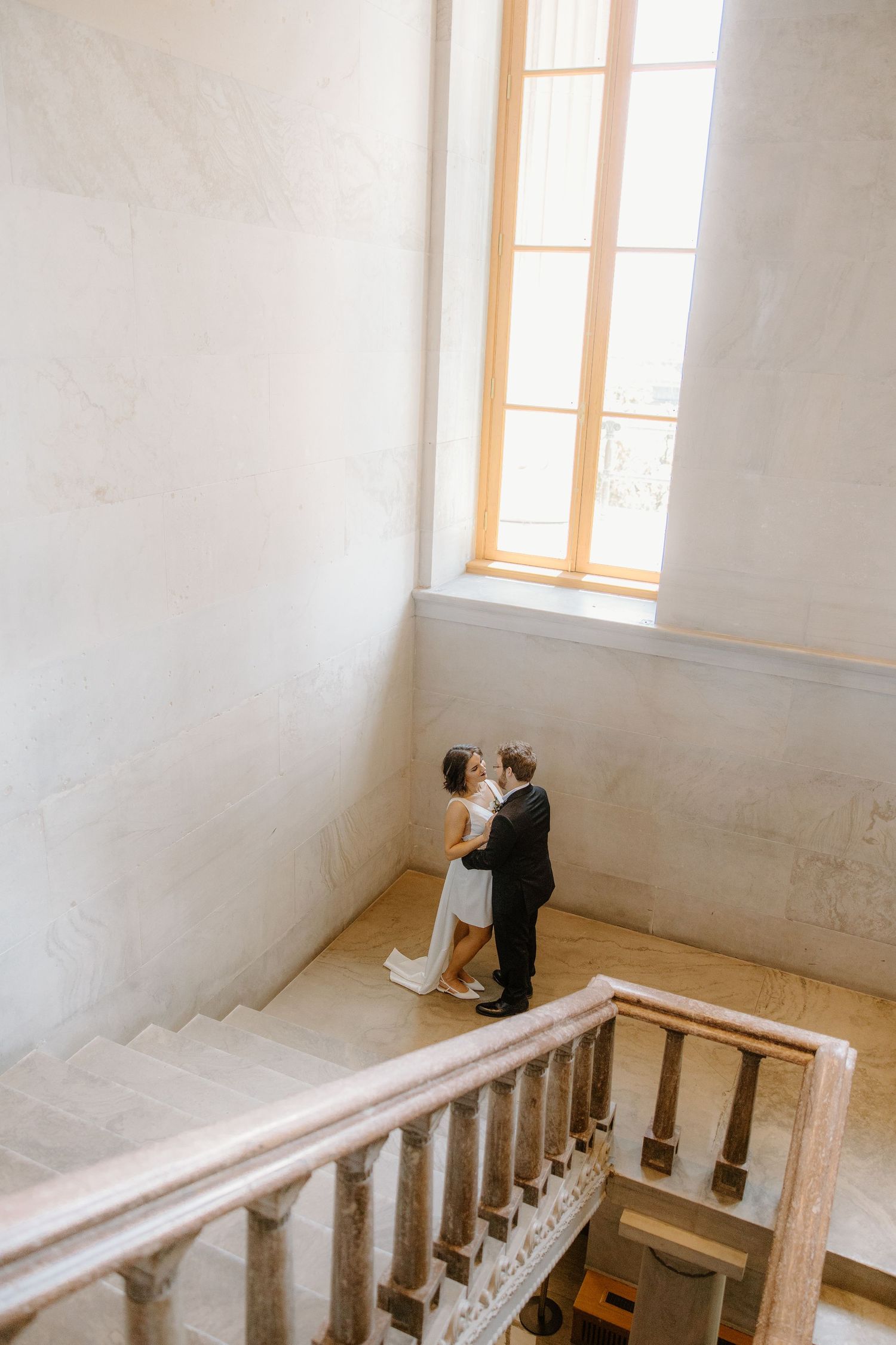 Bride and groom share a romantic moment on marble staircase landing beneath tall window in elegant venue.