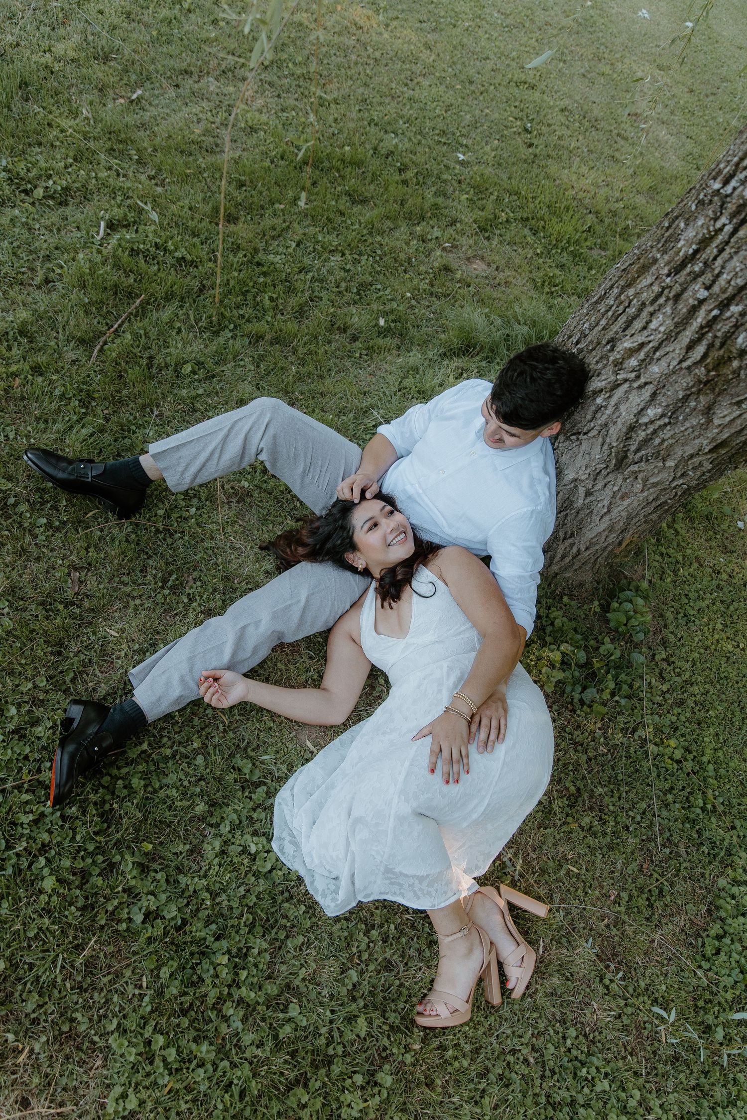 Couple in white outfits relaxing on green grass by a tree trunk in a romantic outdoor setting.