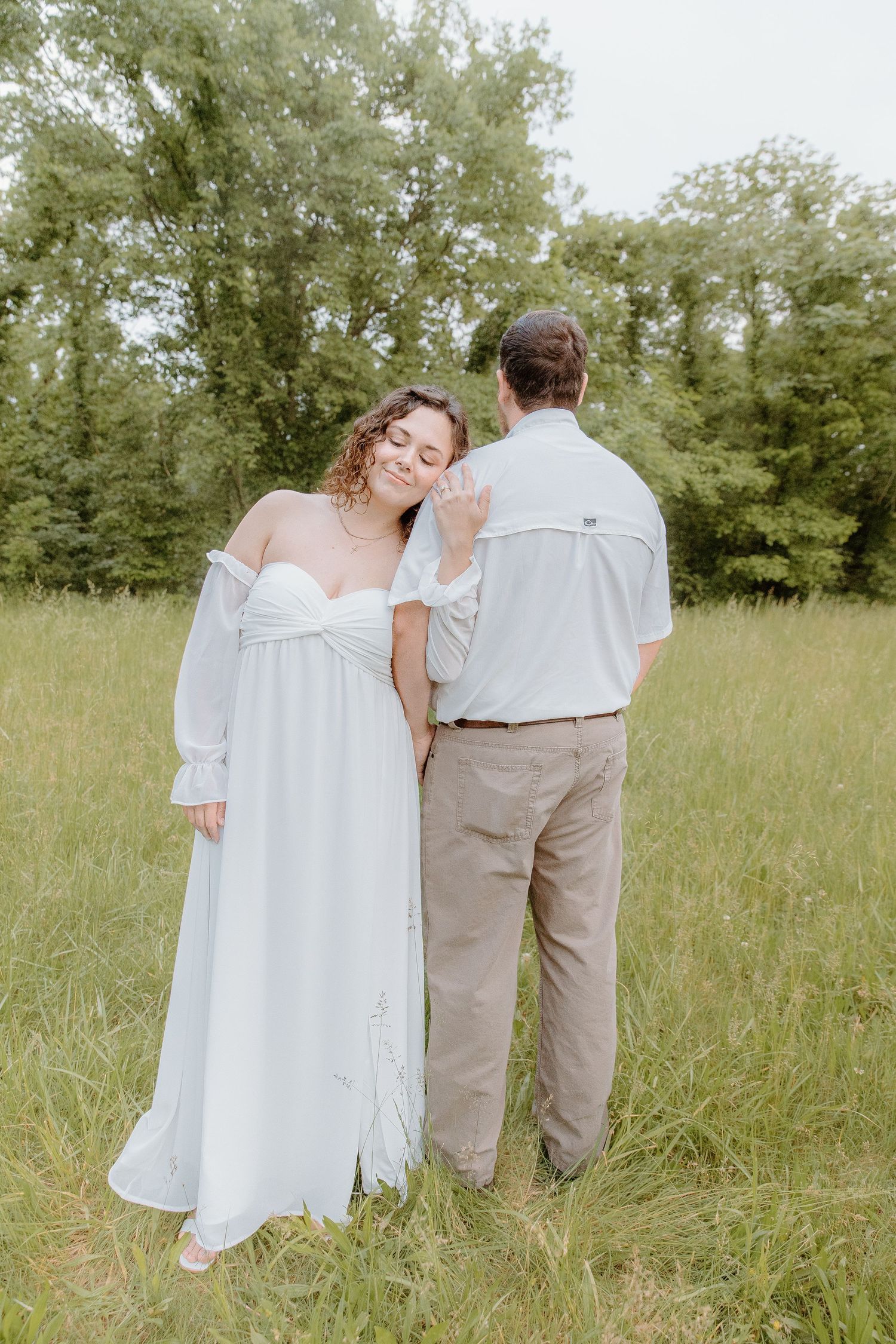 Bride in white off-shoulder gown leans on groom in gray shirt and khaki pants in a lush green meadow.