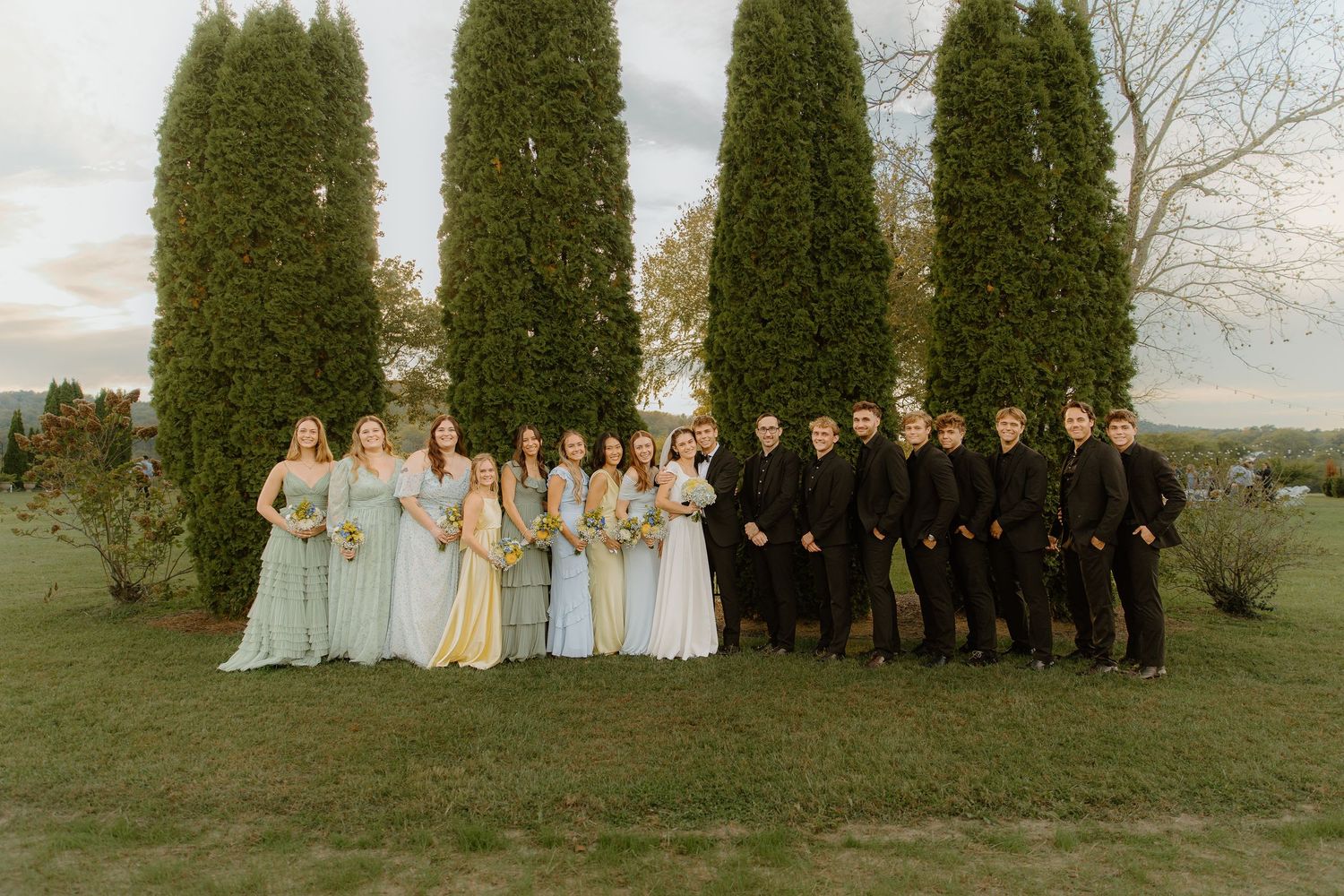 Full wedding party with bridesmaids in mint dresses and groomsmen in black suits by cypress trees.