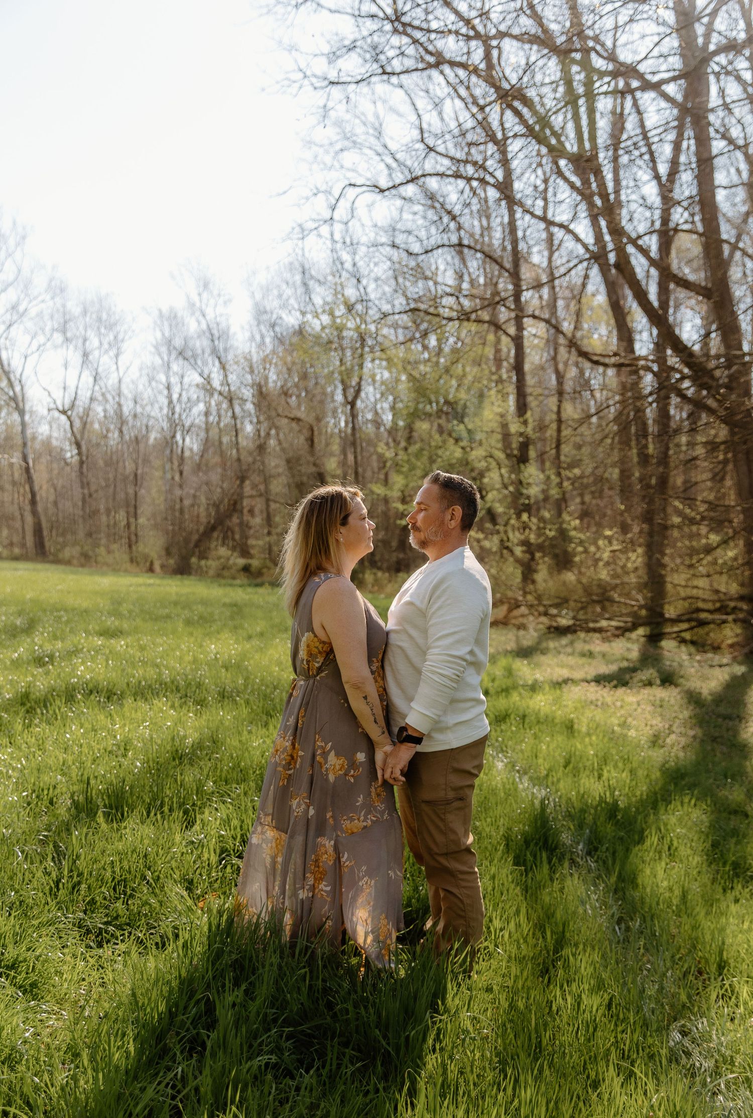 Happy couple holds hands and gazes at each other in a sunny green meadow during engagement session.