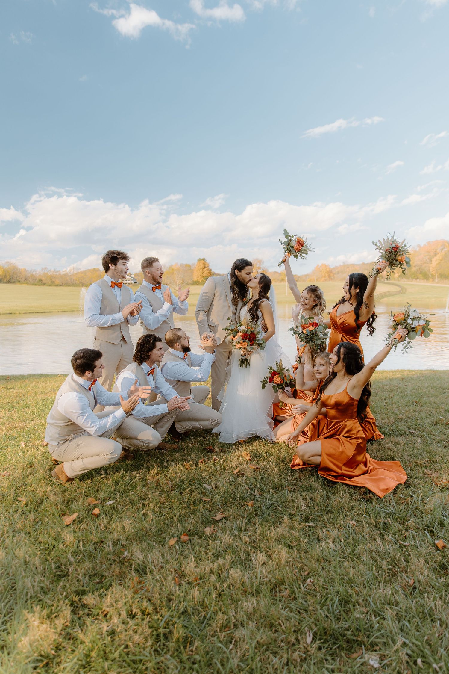Wedding party celebrates outdoors by a lake, bridesmaids in burnt orange dresses as couple shares a kiss.