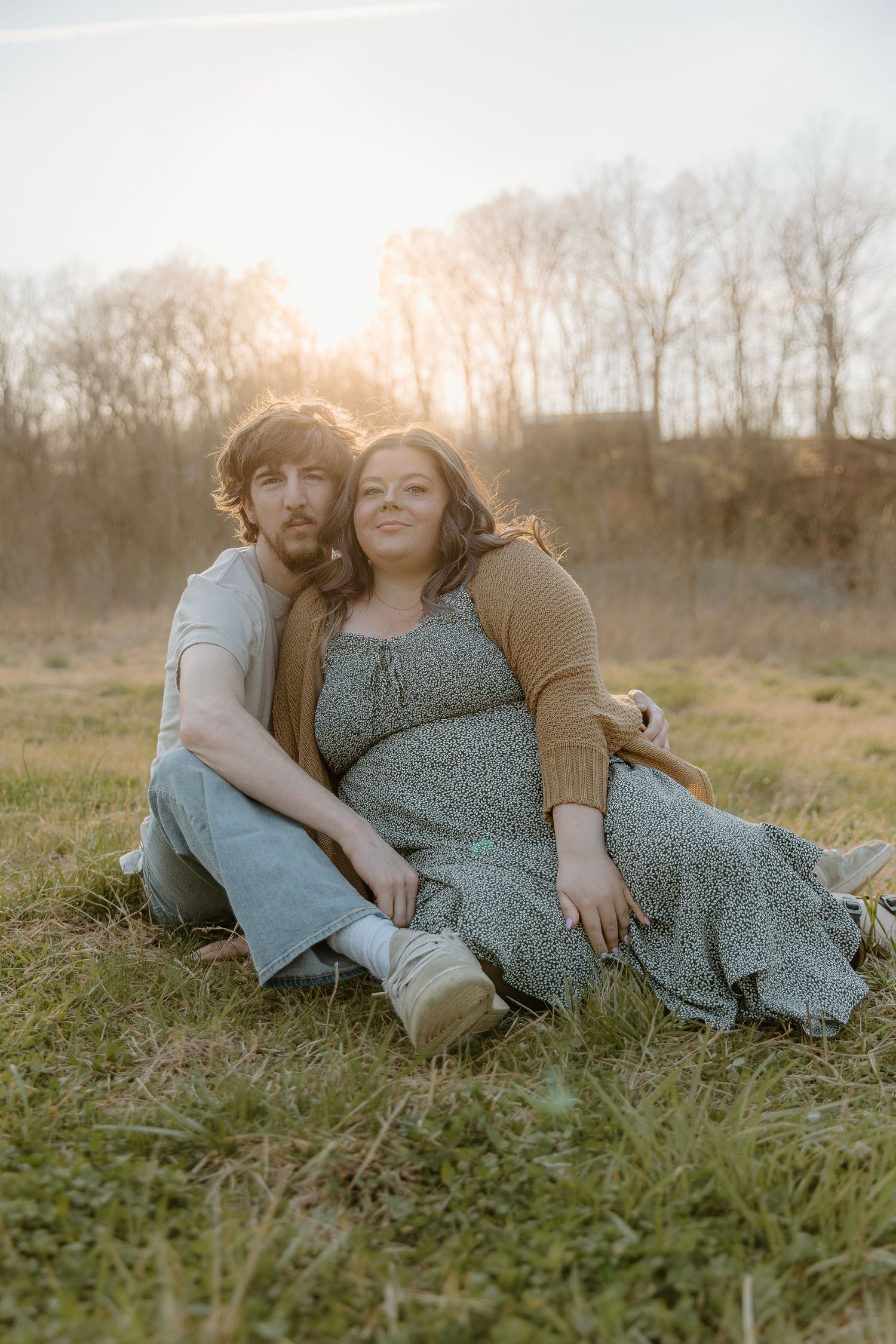 Pregnant couple sits together in a golden sunlit field at sunset during a maternity photo session.