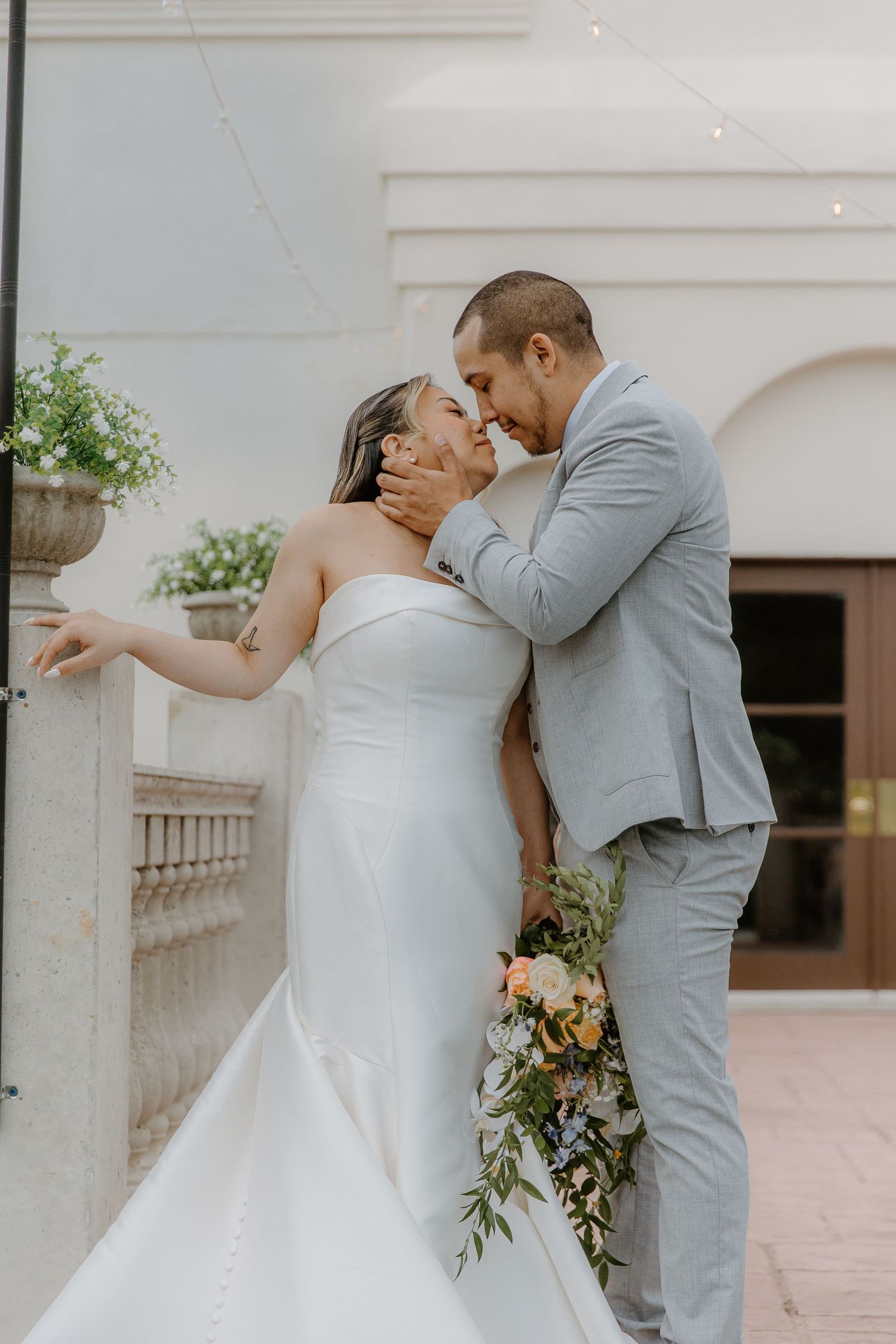 Bride and groom share a romantic kiss outdoors, she in a strapless white gown holding a floral bouquet, he in a light gray suit.