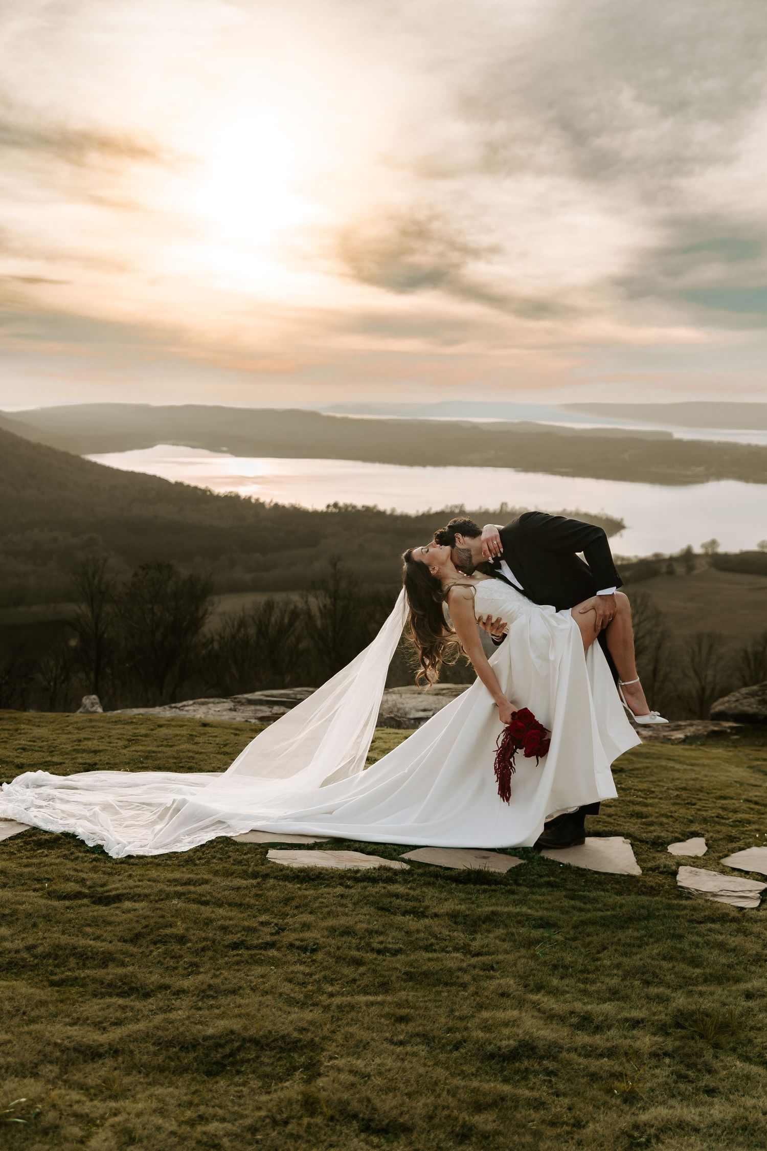 Groom dips bride in a dramatic kiss on a scenic hilltop at sunset with a lake and rolling hills behind them.