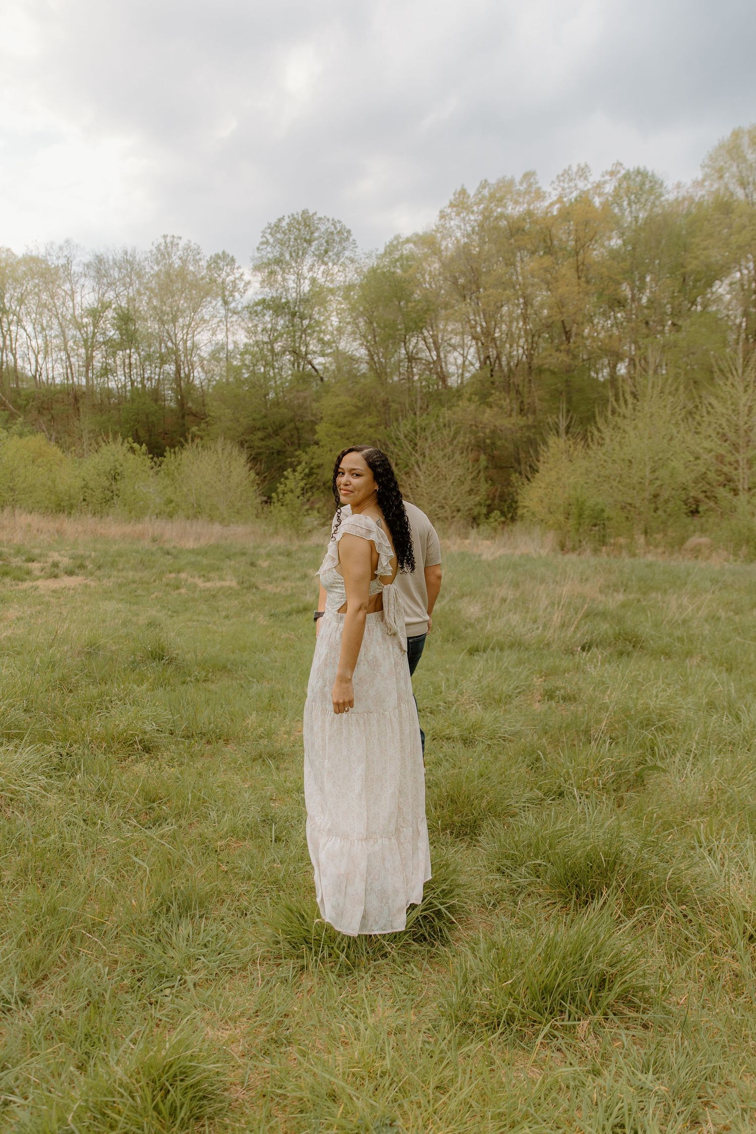 A woman in a white maxi dress stands in a green meadow, smiling over her shoulder, with trees and cloudy sky behind her.