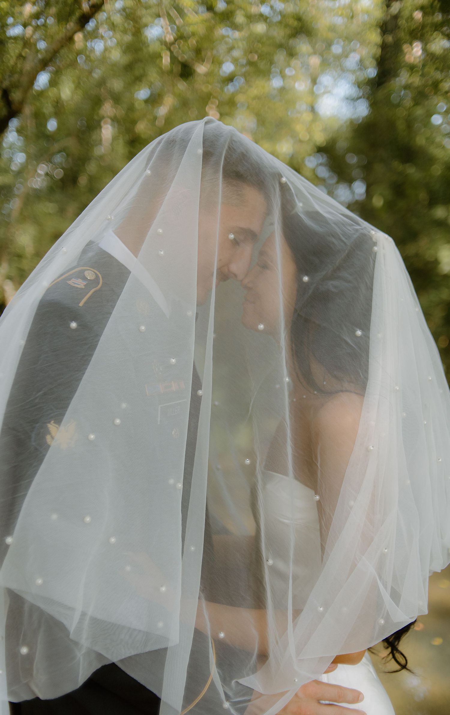 Bride and groom share an intimate moment hidden beneath a polka-dot wedding veil surrounded by greenery.