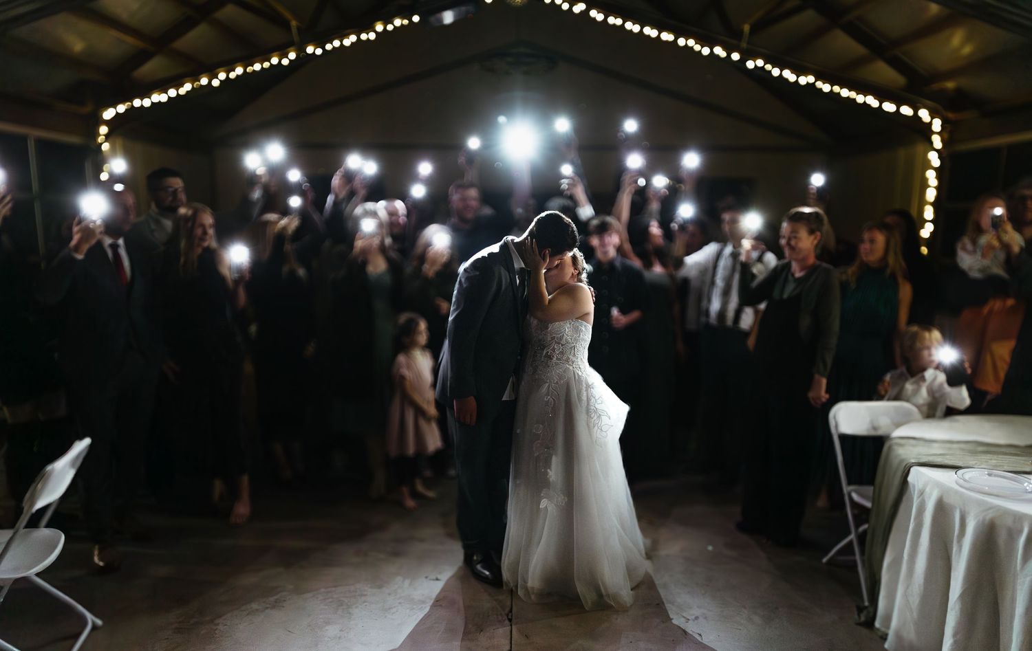 Bride and groom share first dance as wedding guests hold up phone lights in a romantic barn venue.