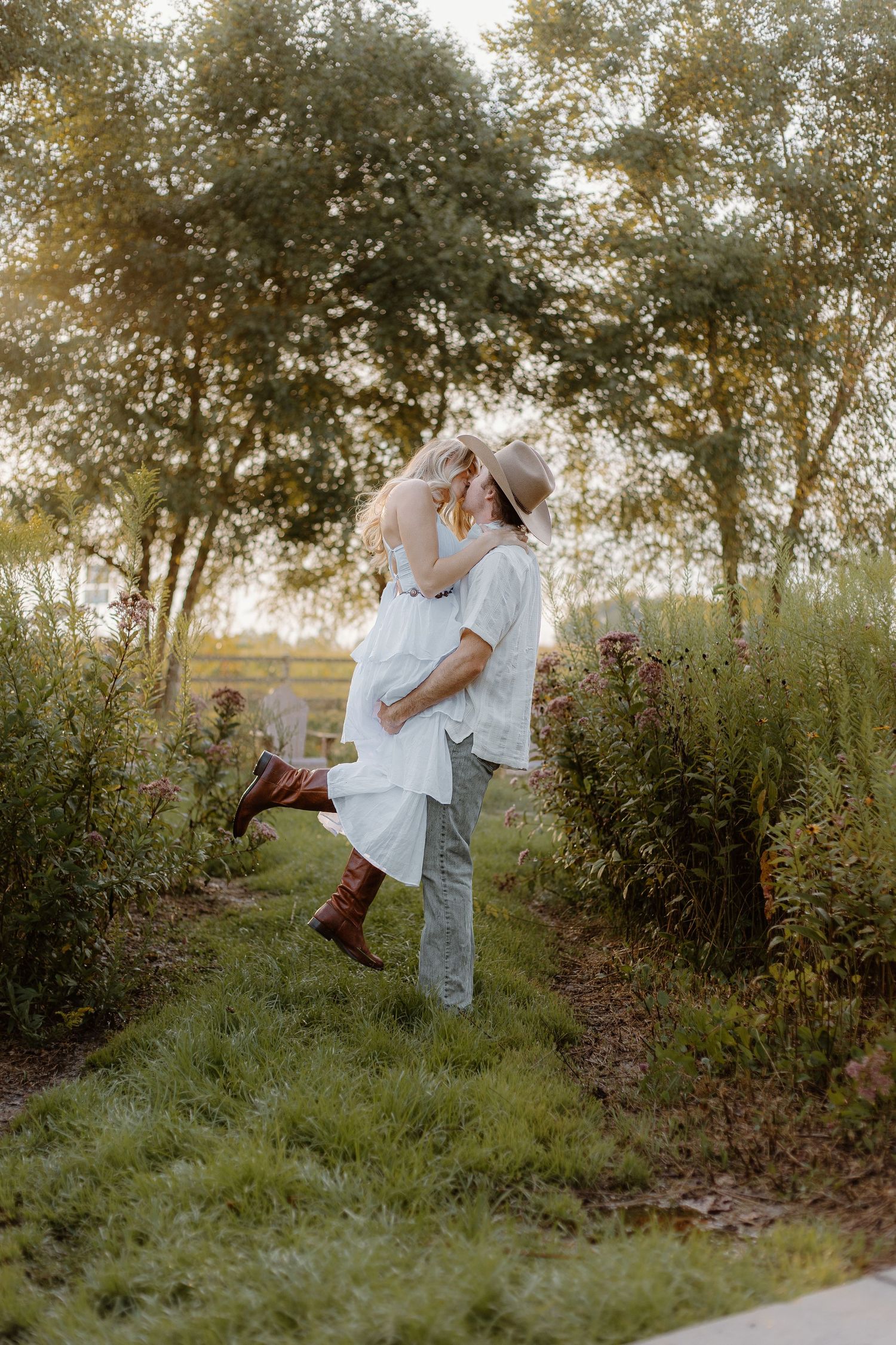 Man lifts and kisses woman in white dress among wildflowers during golden hour engagement session.
