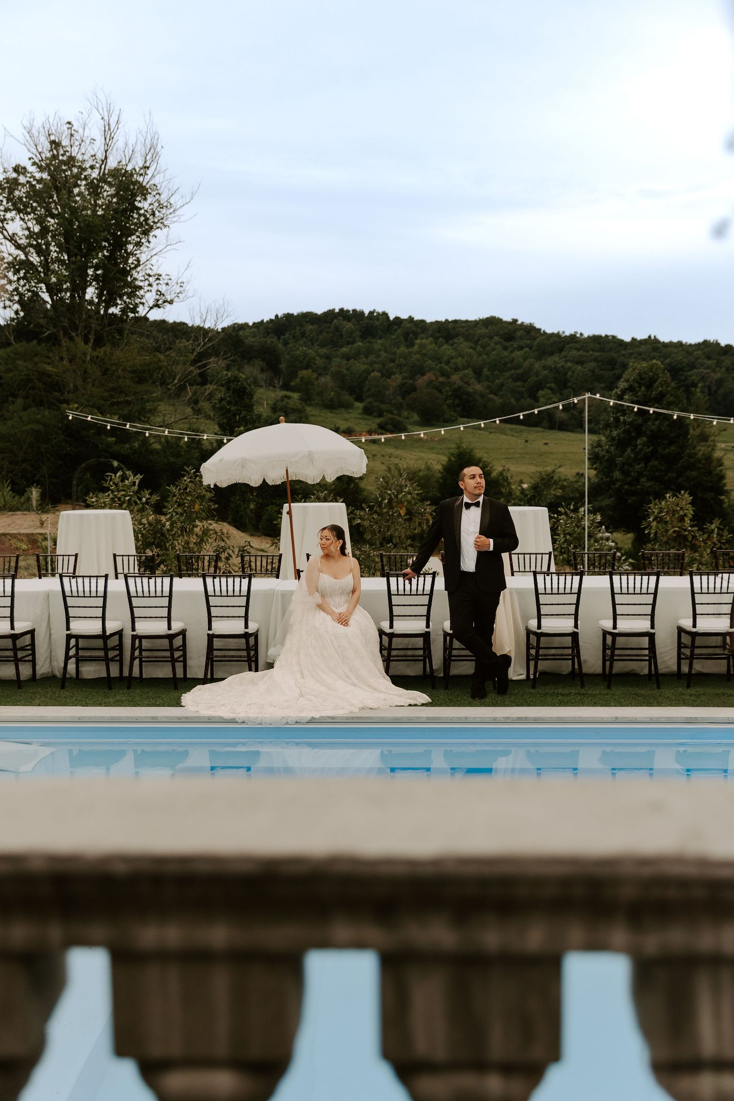 Bride and groom pose poolside at an outdoor wedding reception with rolling green hills.