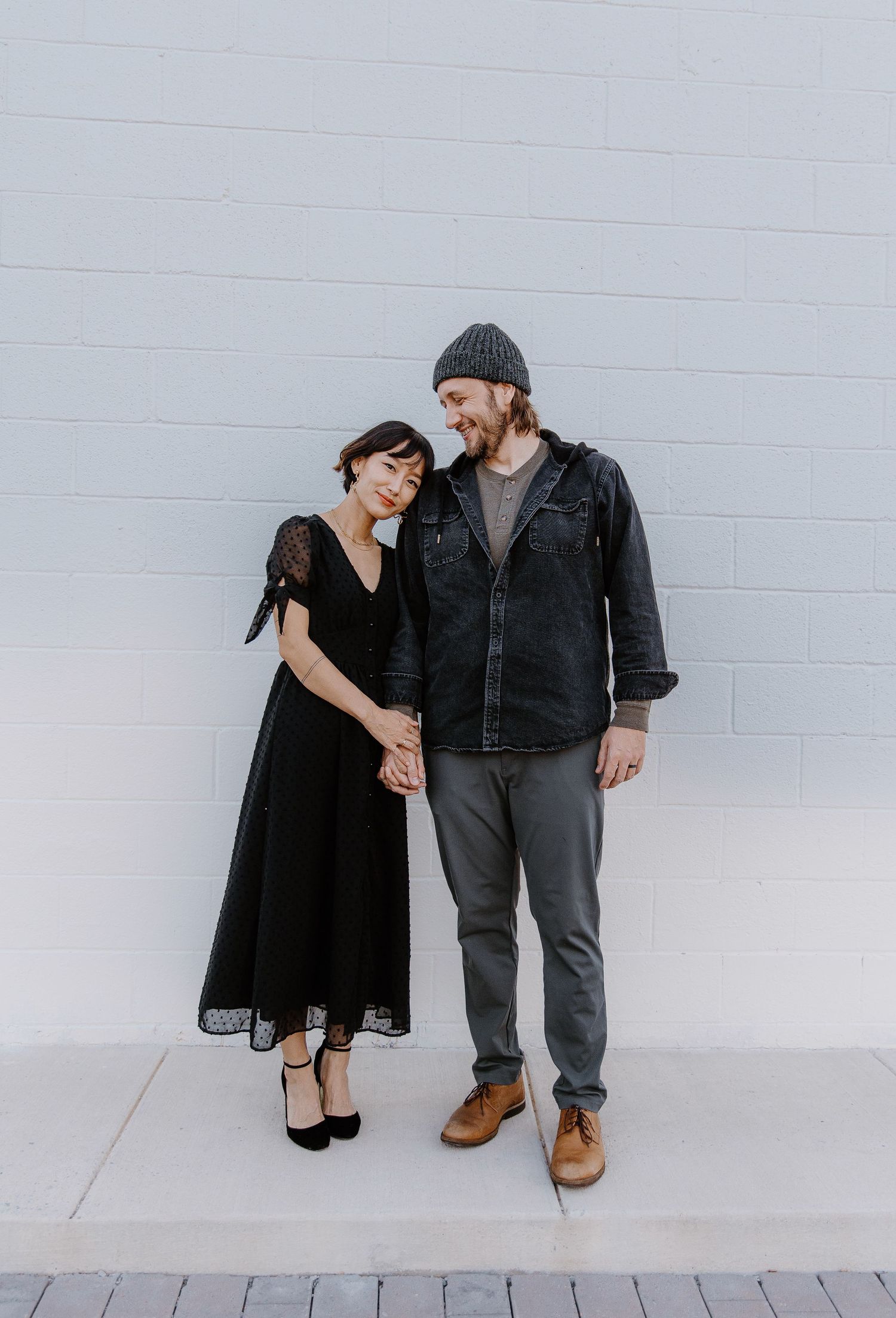 Couple holding hands in front of a white wall, dressed in black.