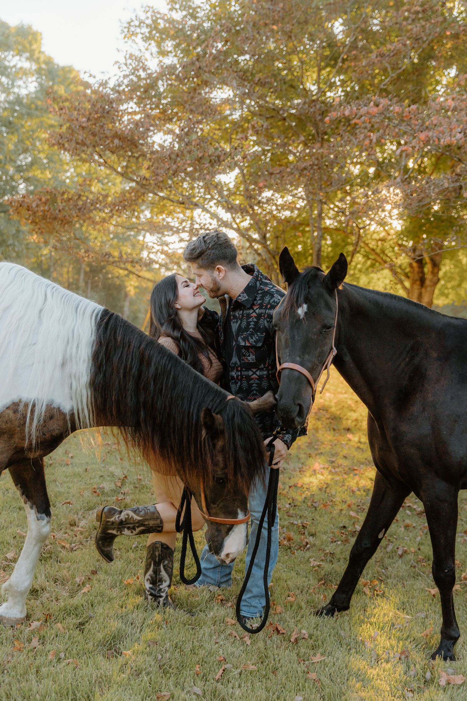 Couple sharing a kiss between two horses in an autumn forest setting.