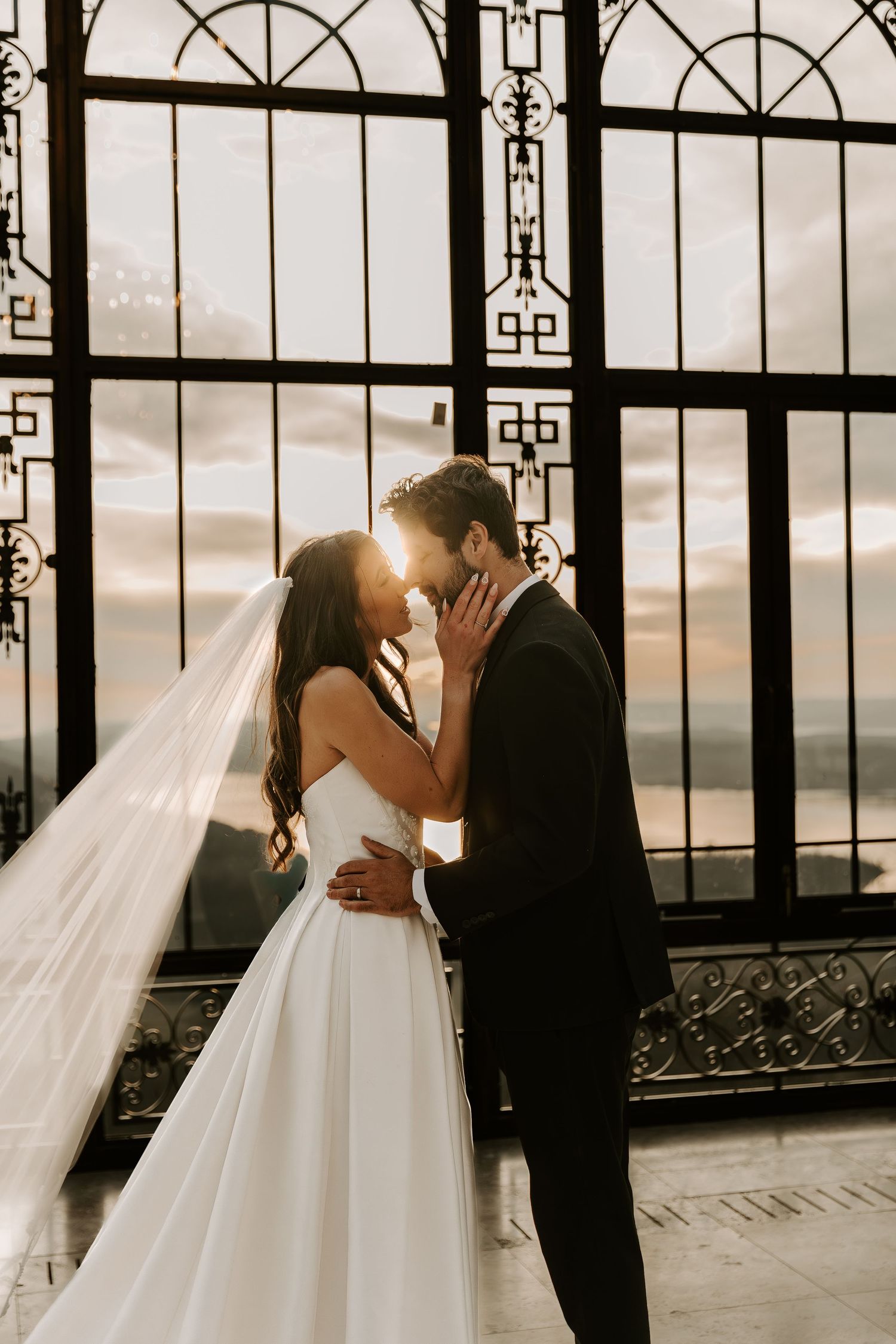 Bride and groom share a romantic kiss at sunset in front of ornate iron gates, bride wearing a flowing white gown and veil.
