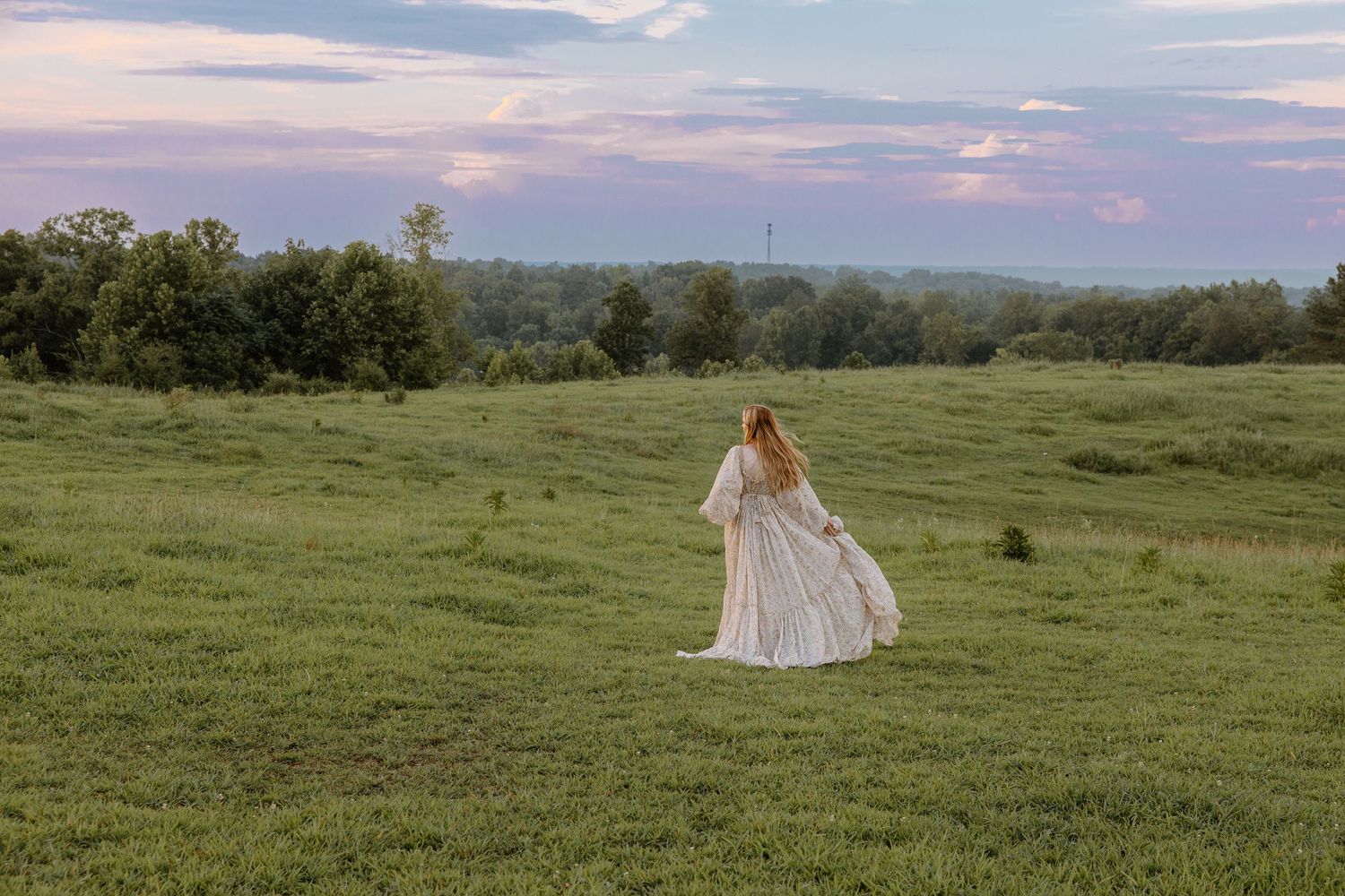 A woman in a flowing white gown walks through a lush green field at dusk with a colorful sky in the background.