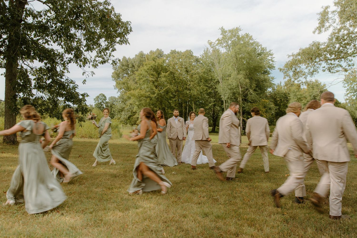 Wedding party in sage green and tan attire running joyfully across a lush green lawn outdoors.