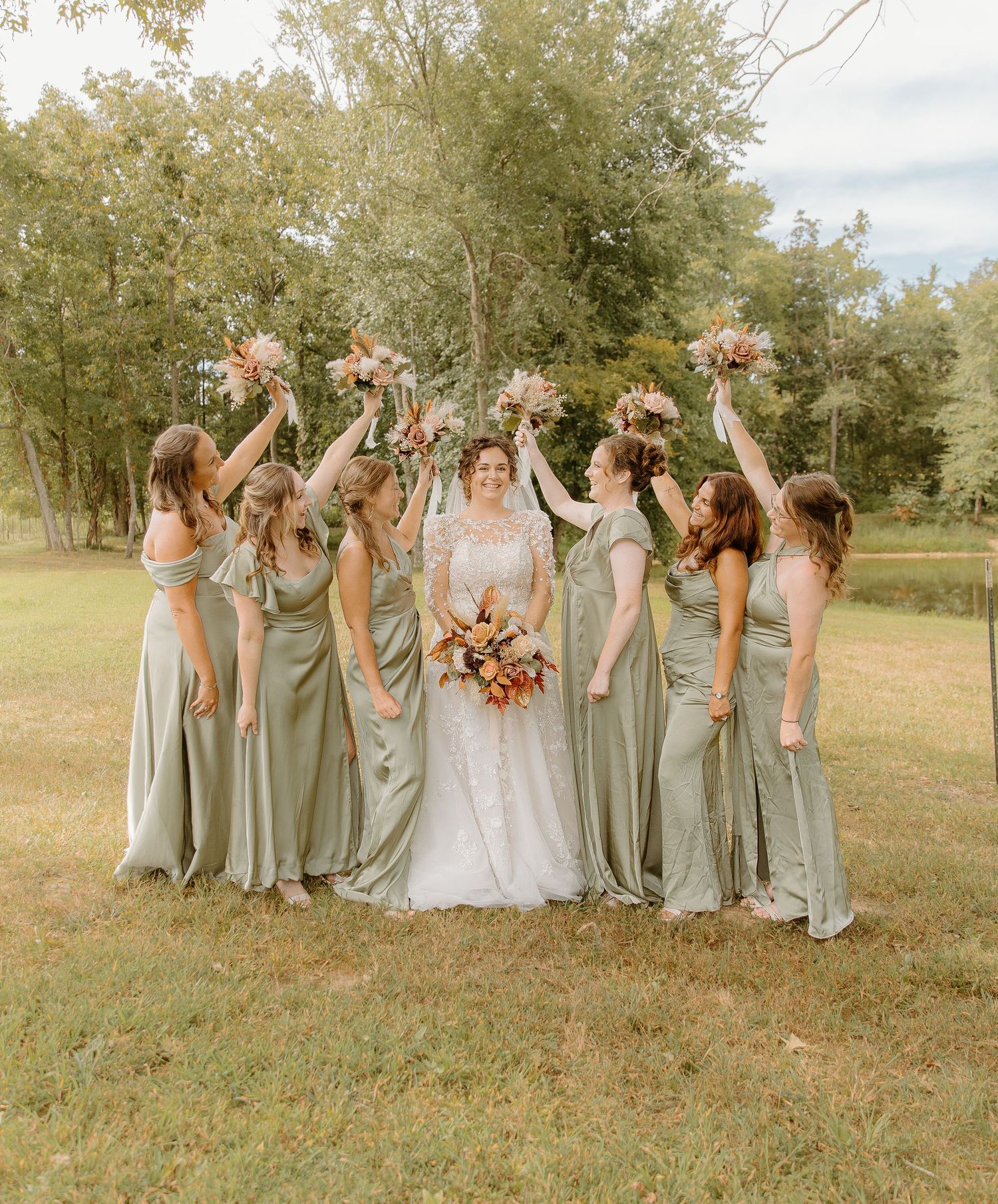 Bride surrounded by bridesmaids in sage green gowns raising bouquets in outdoor woodland setting.