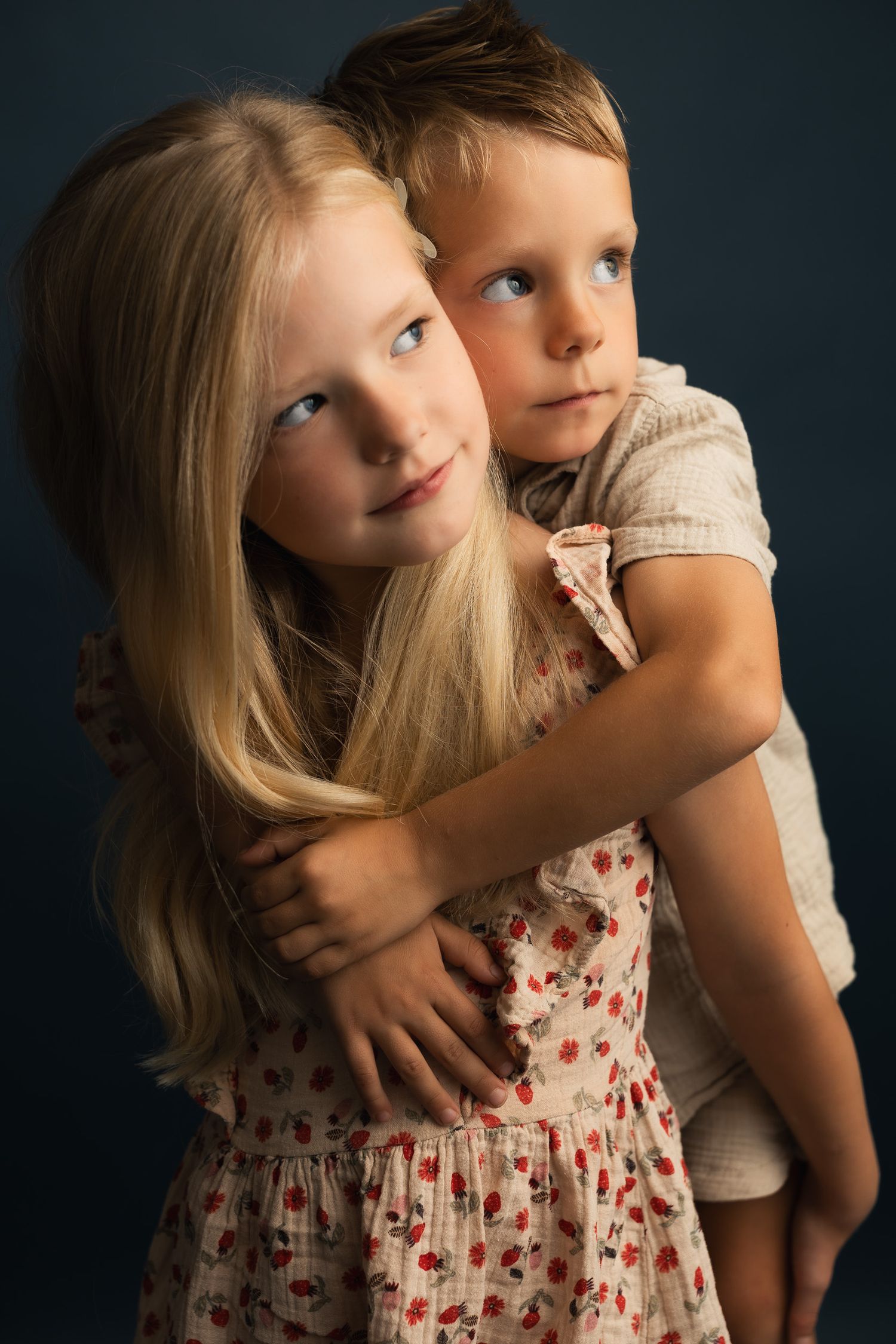 Two blonde children share a warm hug in a studio portrait, girl in floral dress embraced by younger boy.