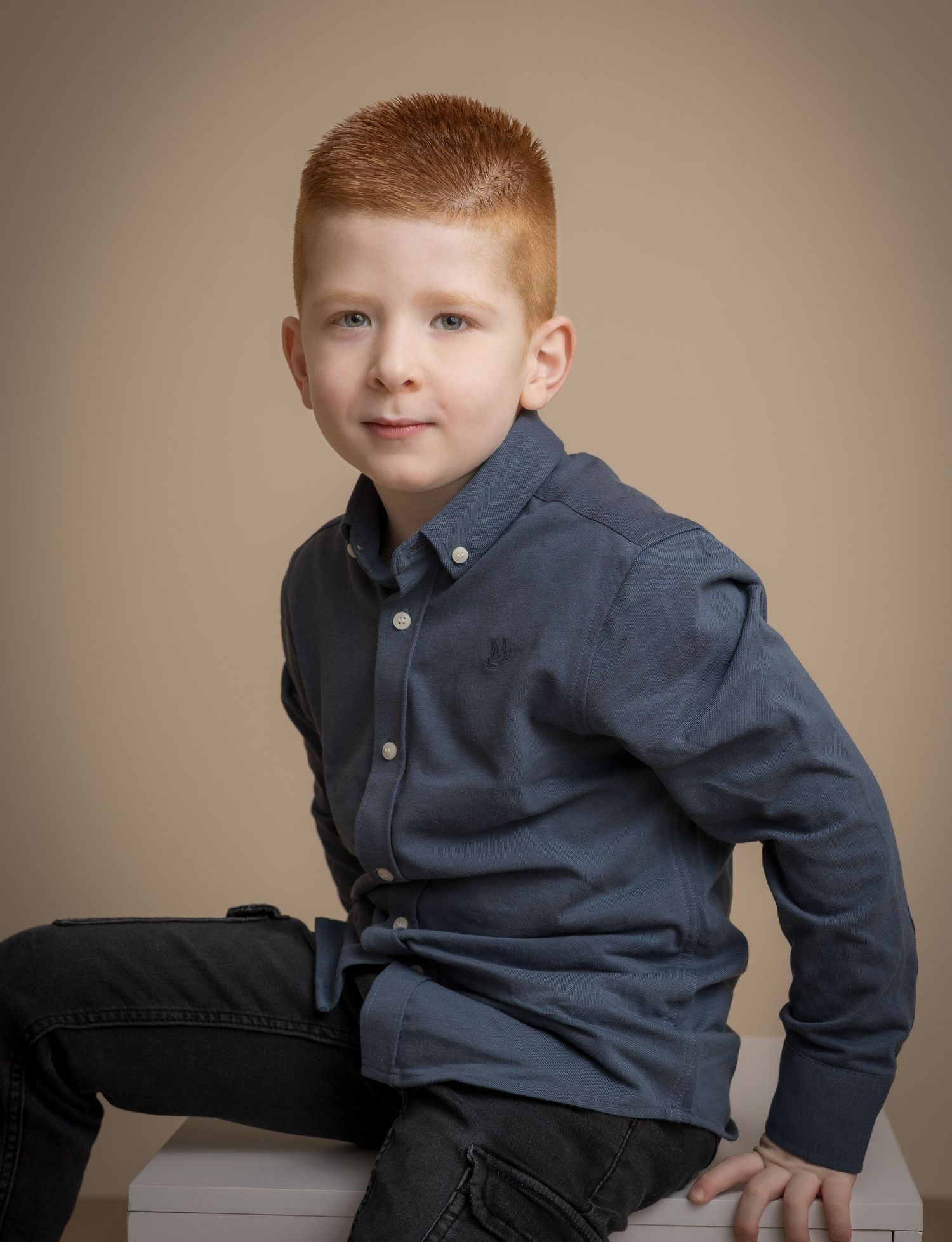 Kinderportret van een roodharige jongen in een blauw overhemd op een witte kubus in de fotostudio.