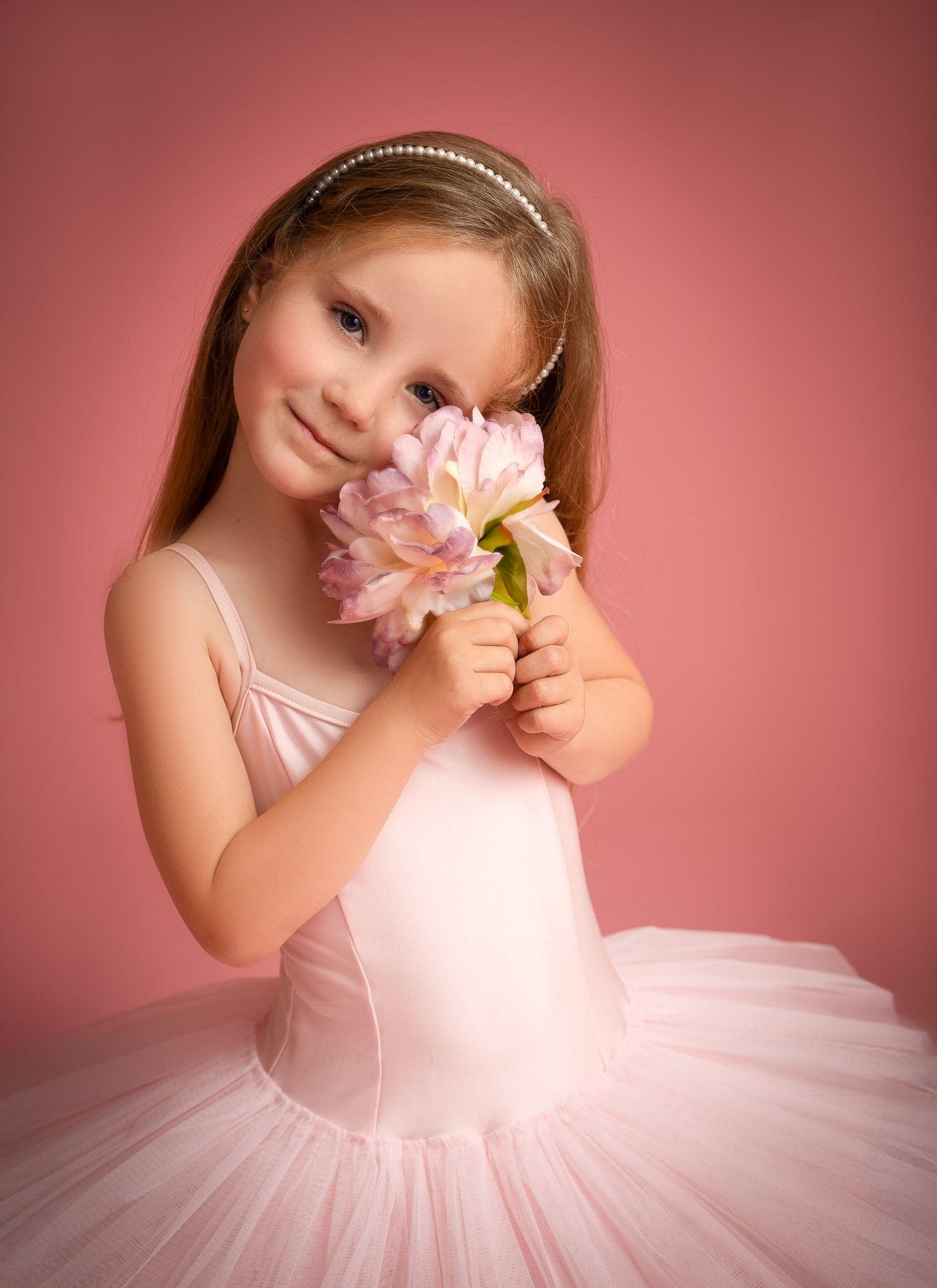 Young girl in pink ballet tutu holds a pink tulip flower, wearing a pearl headband against a pink background.