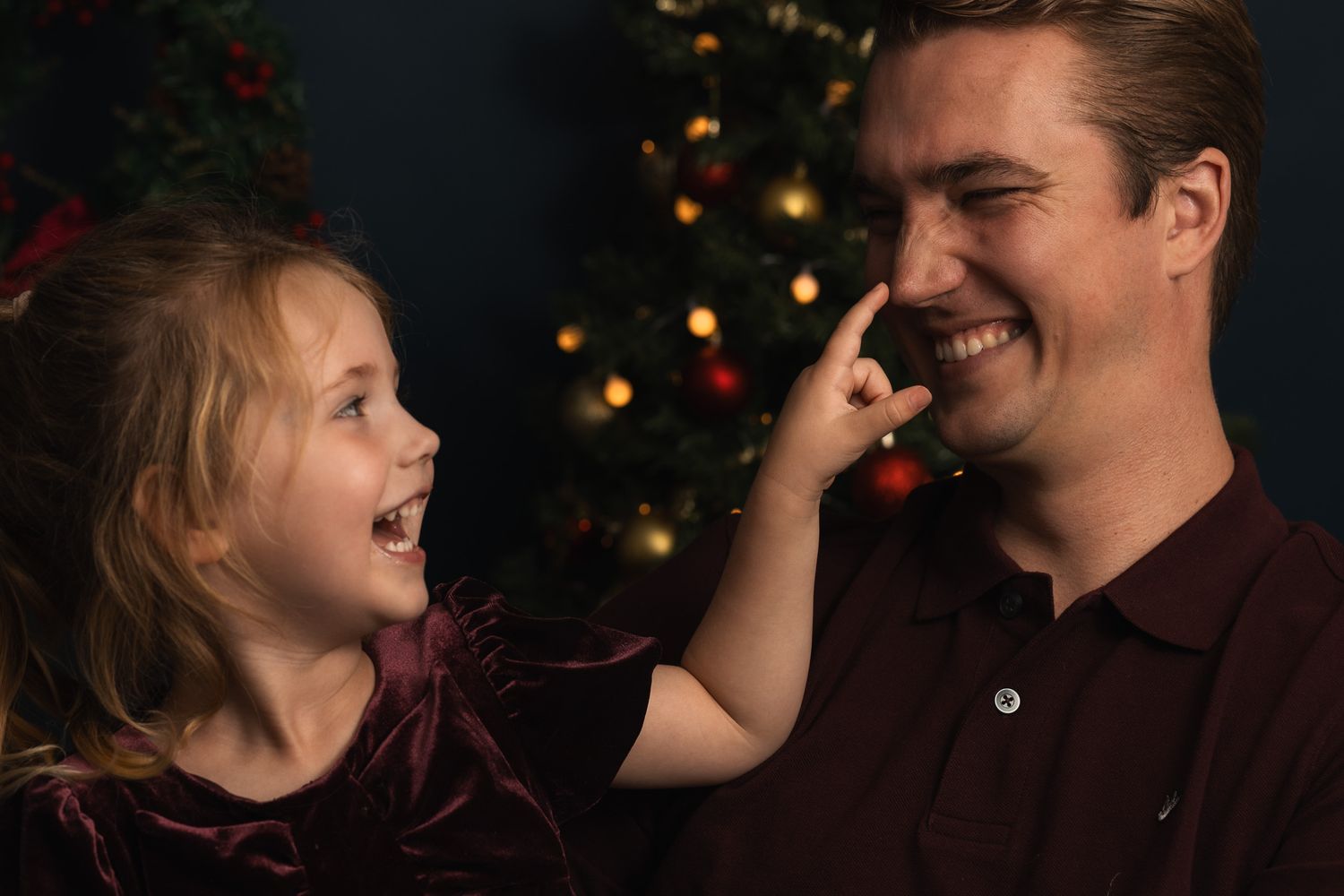 A young girl playfully touches a smiling man's nose in front of a Christmas tree during a festive holiday portrait.